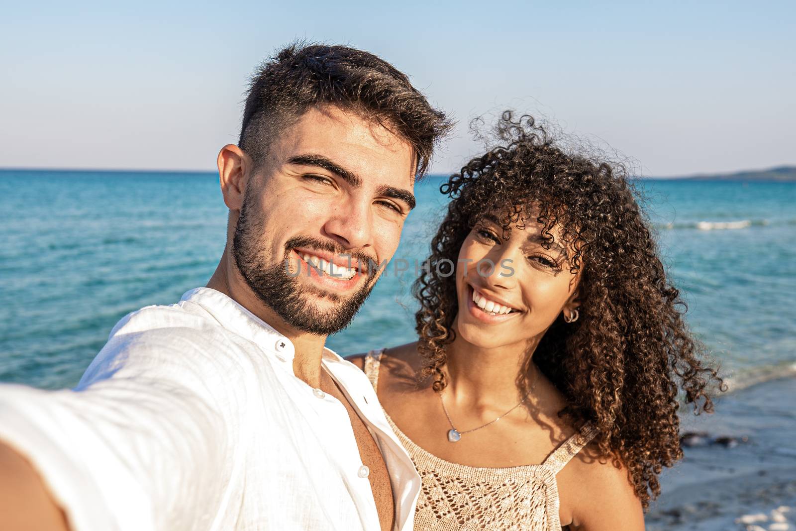 Beautiful mixed race couple taking a selfie at the sea at dusk - Handsome young man in white open shirt with his latina curly haired girlfriend smiling together in summer vacation by robbyfontanesi