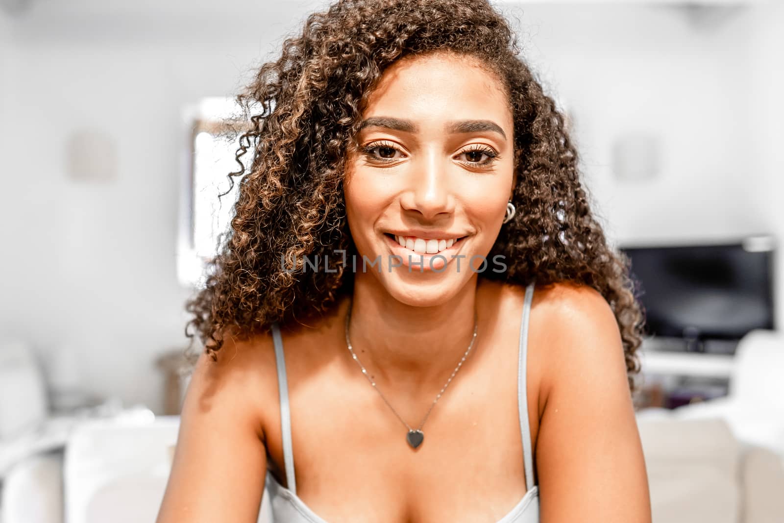 Close-up of young hispanic cute curly woman smiling looking at the camera like as in video conference in a white home living room ambient - Female confident millennial latina portrait in singlet vest by robbyfontanesi