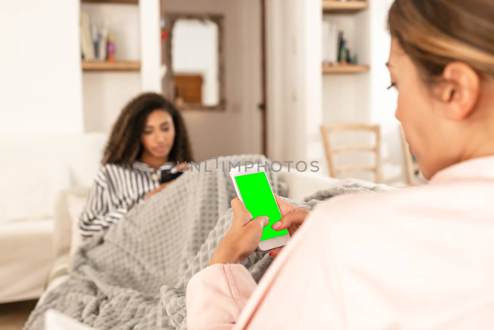 Two young mixed race female friends always connected communicating wearing pajama snuggle up on the sofa under a blanket - Selective focus on the green screen of the white smartphone by robbyfontanesi