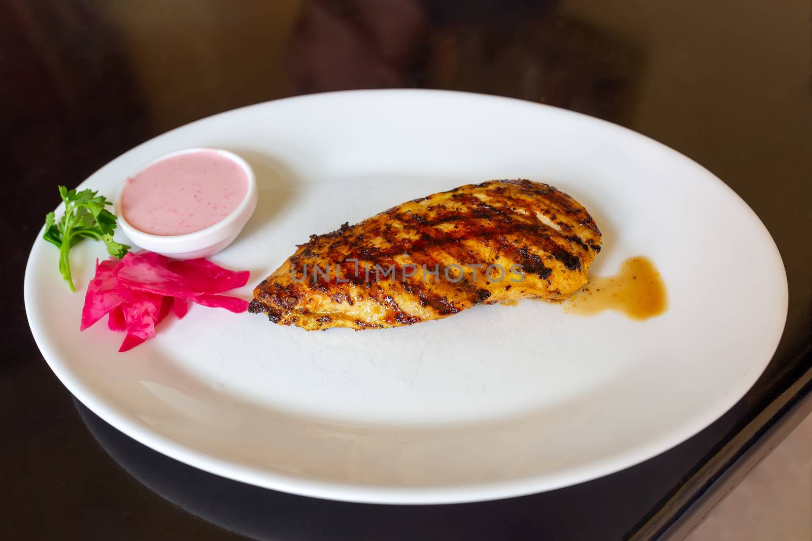 Appetizing grilled chicken breast on a white plate with pink sauce, pickled ginger and parsley. Nearby lies a fork and a knife in a napkin.