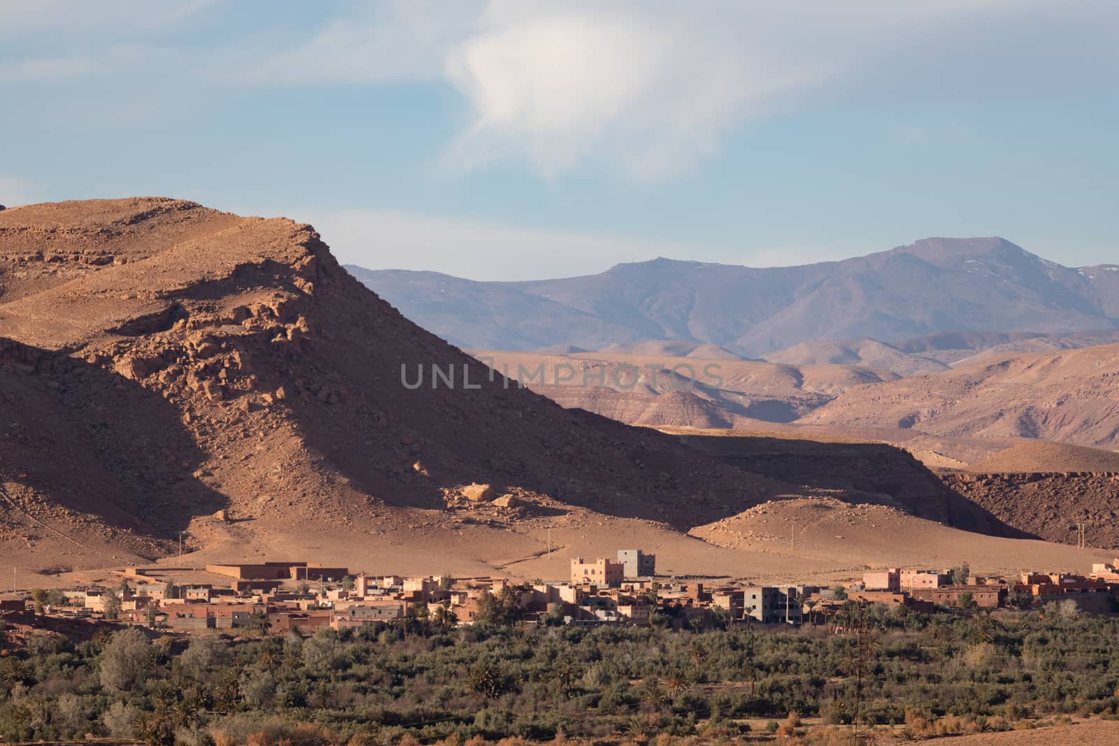 Atlas mountains with town seen from Ait Ben Haddou ksar Morocco, ancient fortress that is a Unesco Heritage site. Beautiful late afternoon light with honey, gold coloured mud brick construction the kasbah, or fortified town dates from 11th cent. and is on the former caravan route from the Sahara and Marrakech. The location has been used for many famous movies. High quality photo