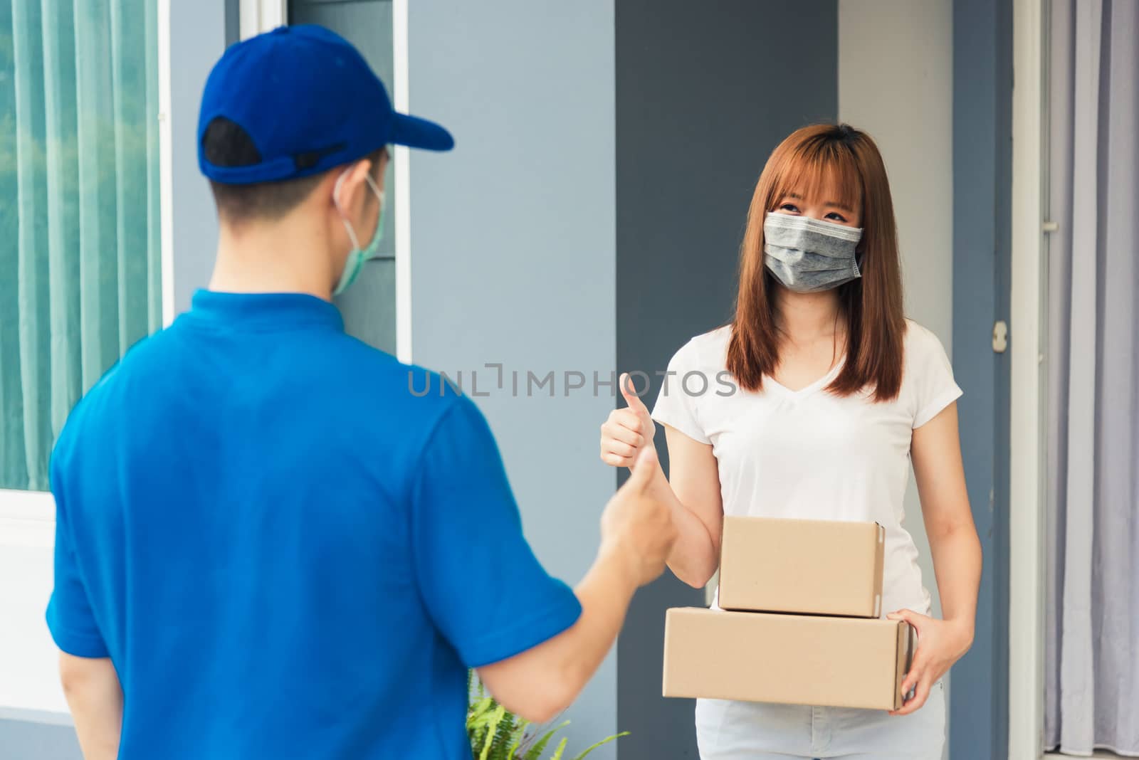 Asian delivery express courier young man giving parcel boxes to woman customer receiving both protective face mask and show thumbs up finger for good support sign, under curfew pandemic coronavirus