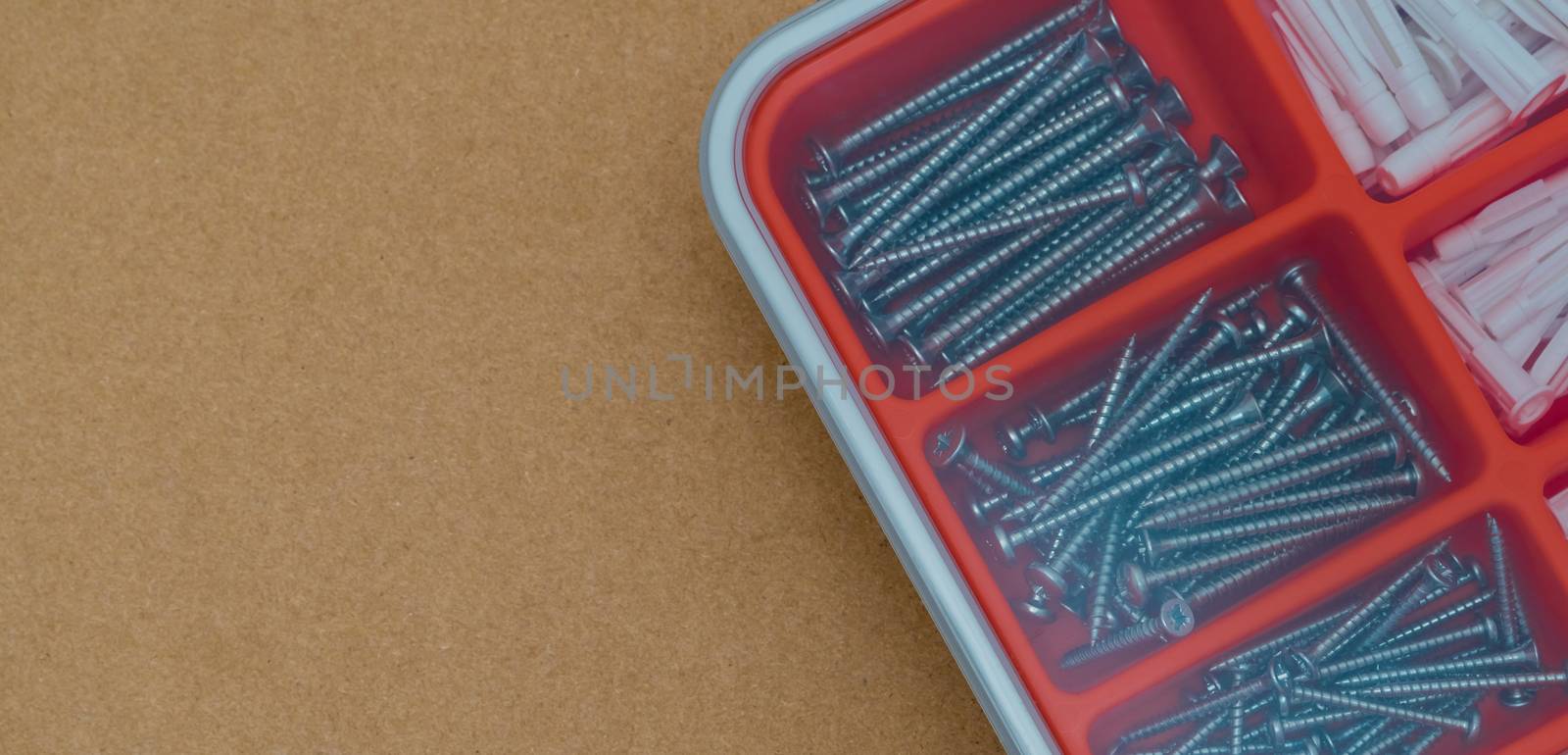 Steel silver screws and wall plug in orange plastic box. Top view tool box on brown paper box background with copy space. Set of screws and plastic plug for drilling wall and installation things.
