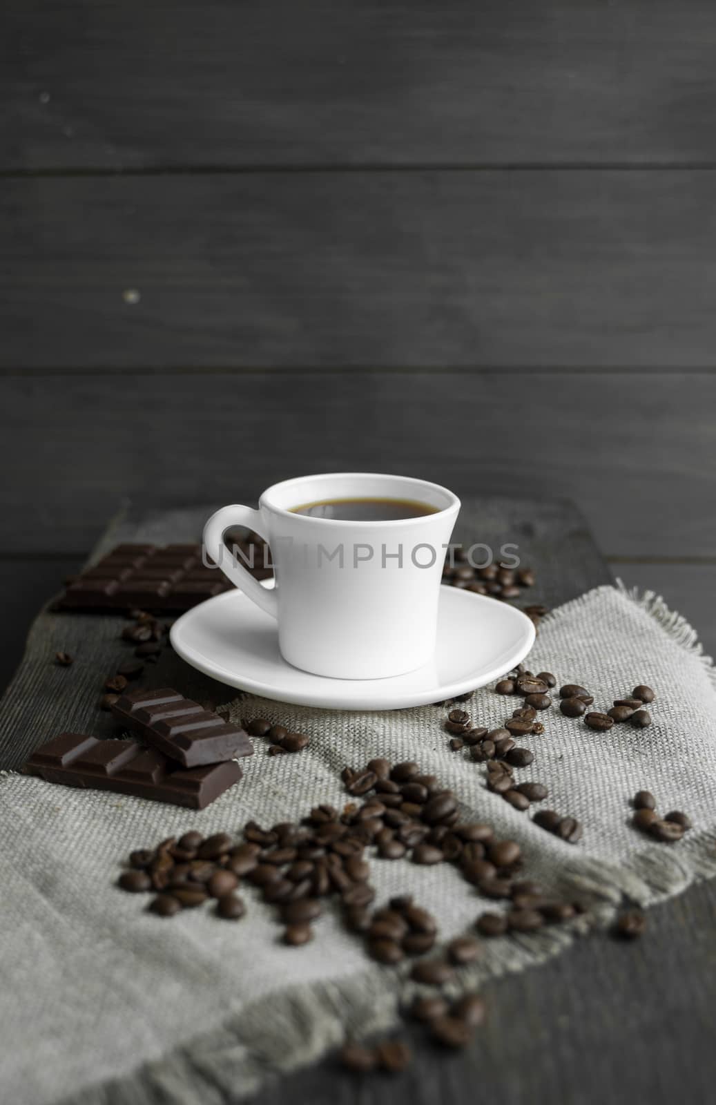 Coffee cup with cookies and chocolate with scattered coffee beans on linen and wooden table background. Mug of black coffee