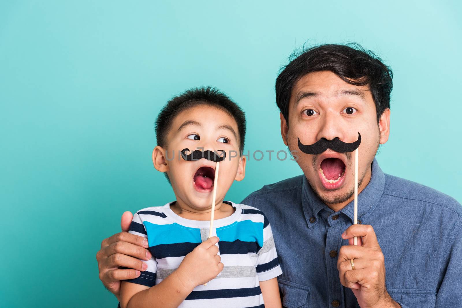 Family funny happy hipster father and his son kid holding black mustache props for the photo booth close face, studio shot isolated on a blue background, November men health awareness