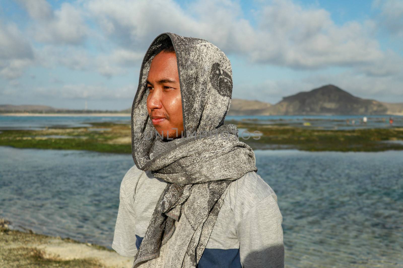 Close up teenage boy with towel on the head in casual clothes. A Guy on the beach. The guy in the towel at sea. Young asian man looks at the landscape.
