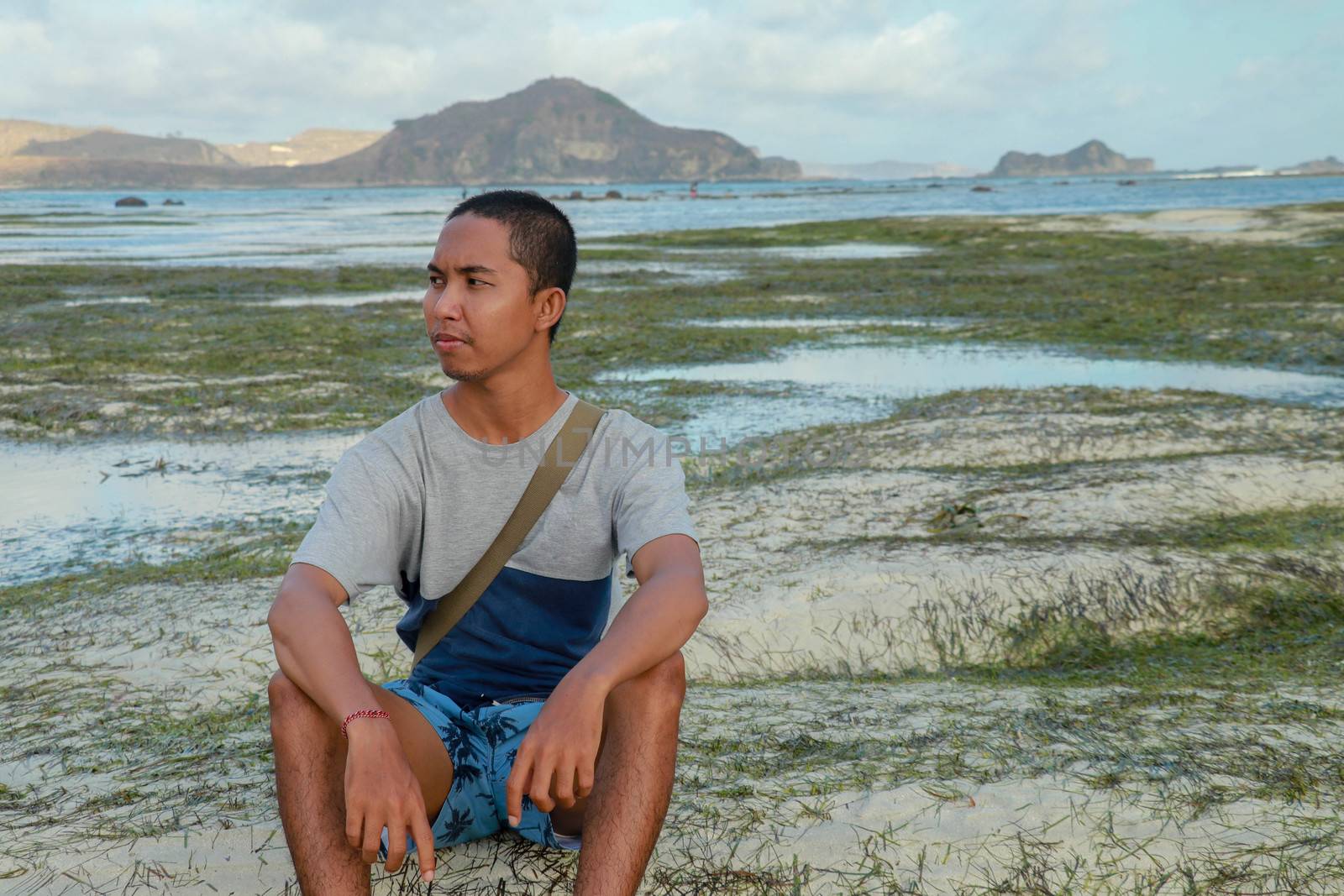 Man thinking and watching the sea on the beach. Teenager sitting on white sand at Tanjung Aan beach, Lombok, Indonesia. A young Asian man looks at the landscape.