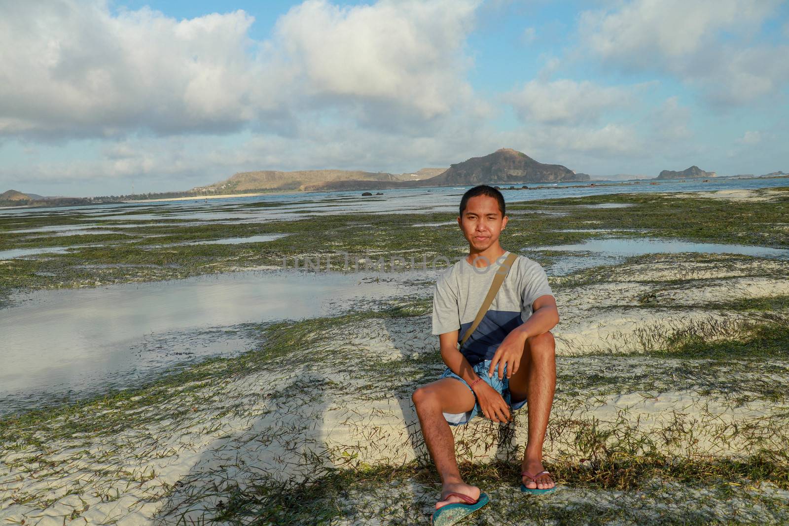 Man thinking and watching the sea on the beach. Teenager sitting on white sand at Tanjung Aan beach, Lombok, Indonesia. A young Asian man looks at the landscape.