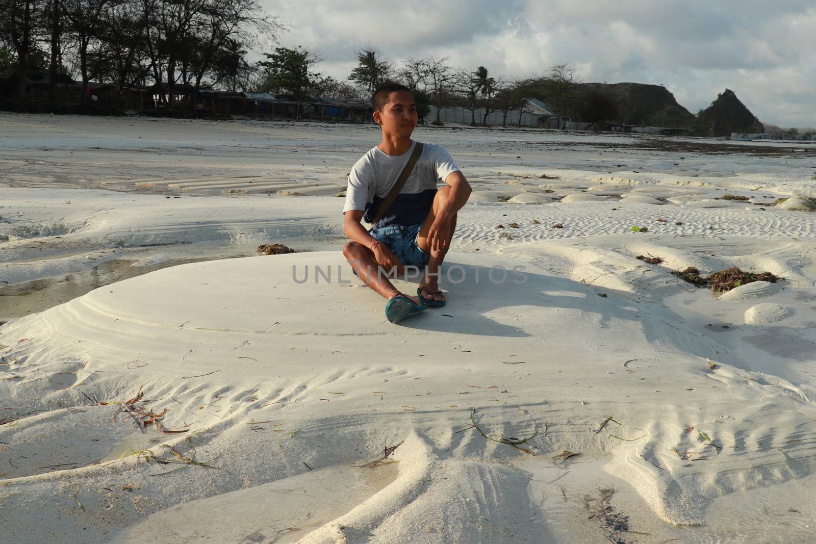 Man thinking and watching the sea on the beach. Teenager sitting on white sand at Tanjung Aan beach, Lombok, Indonesia. A young Asian man looks at the landscape by Sanatana2008