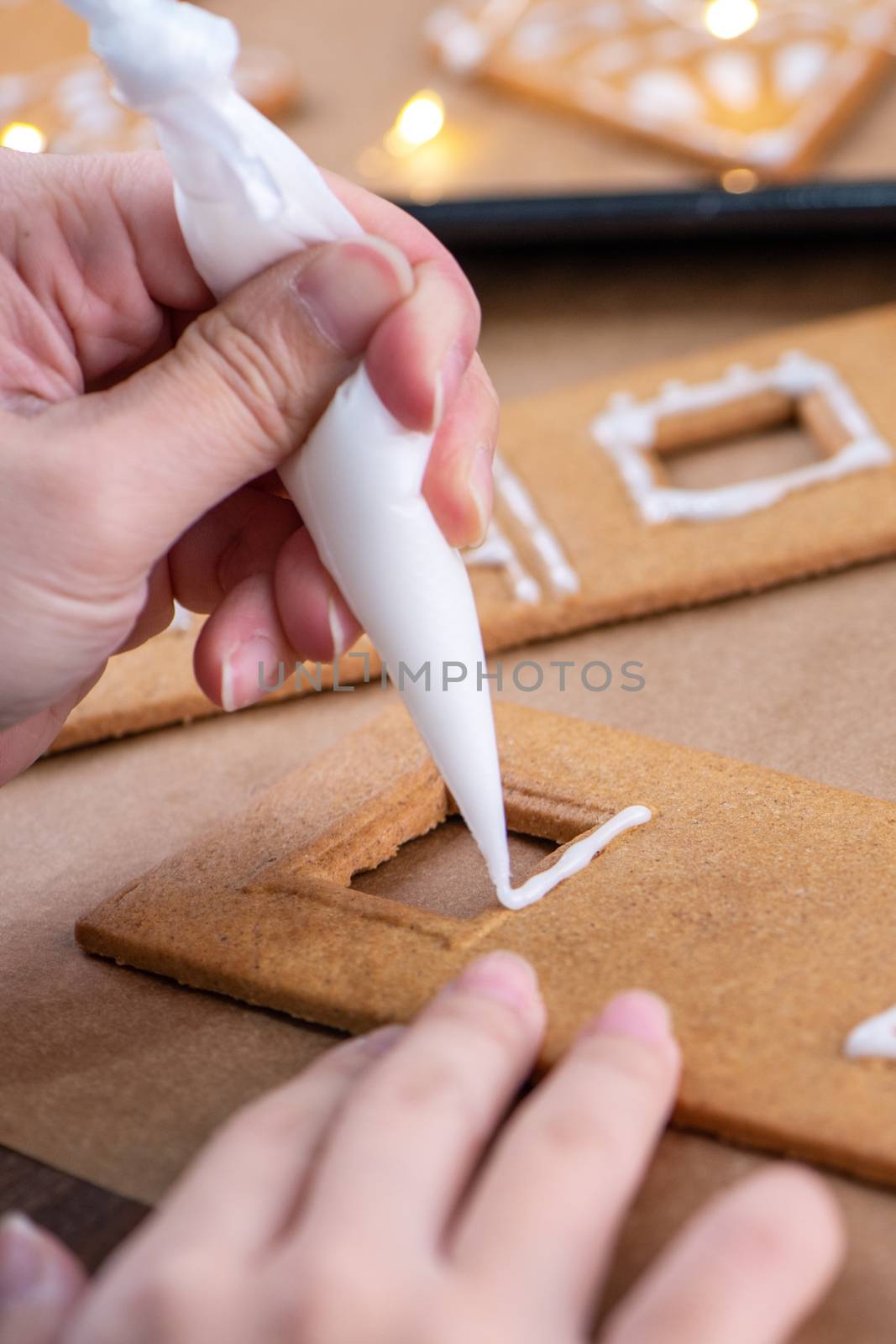 Young woman is decorating Christmas Gingerbread House cookies biscuit at home with frosting topping in icing bag, close up, lifestyle.