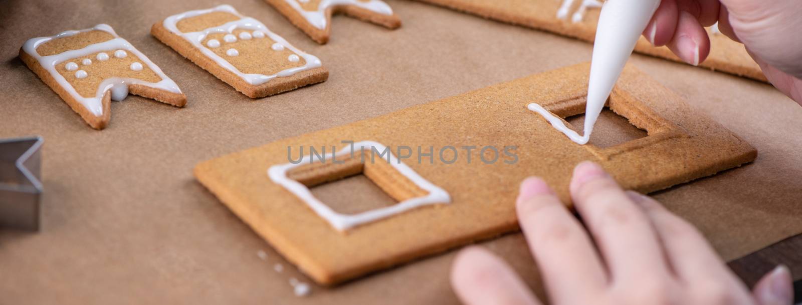 Young woman is decorating Christmas Gingerbread House cookies biscuit at home with frosting topping in icing bag, close up, lifestyle.