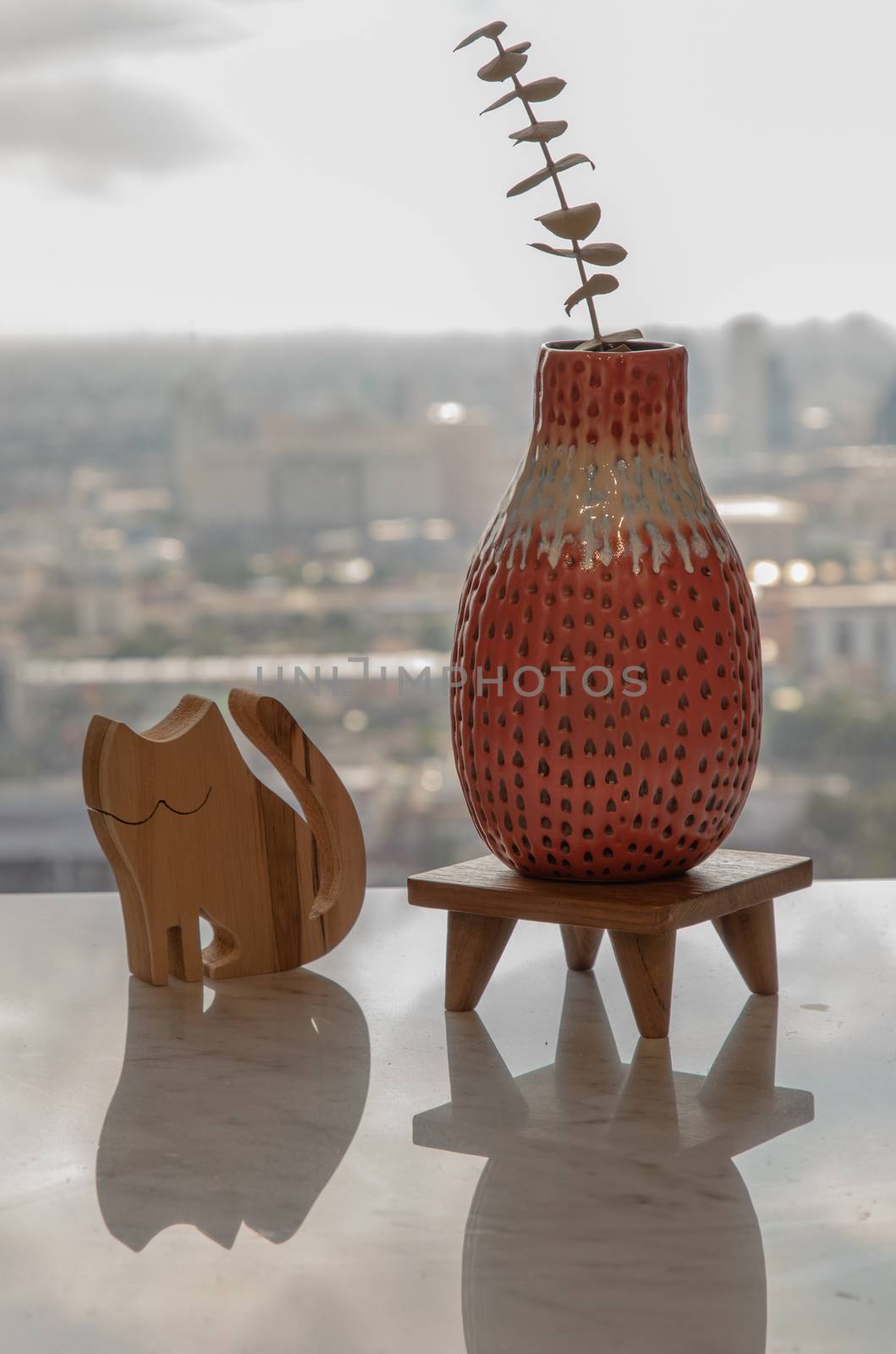 Dried flowers in beautiful ceramic red vase handmade with wooden doll in cat shape on marble table of living room and city view. Home decor. Space for text. Selective focus.