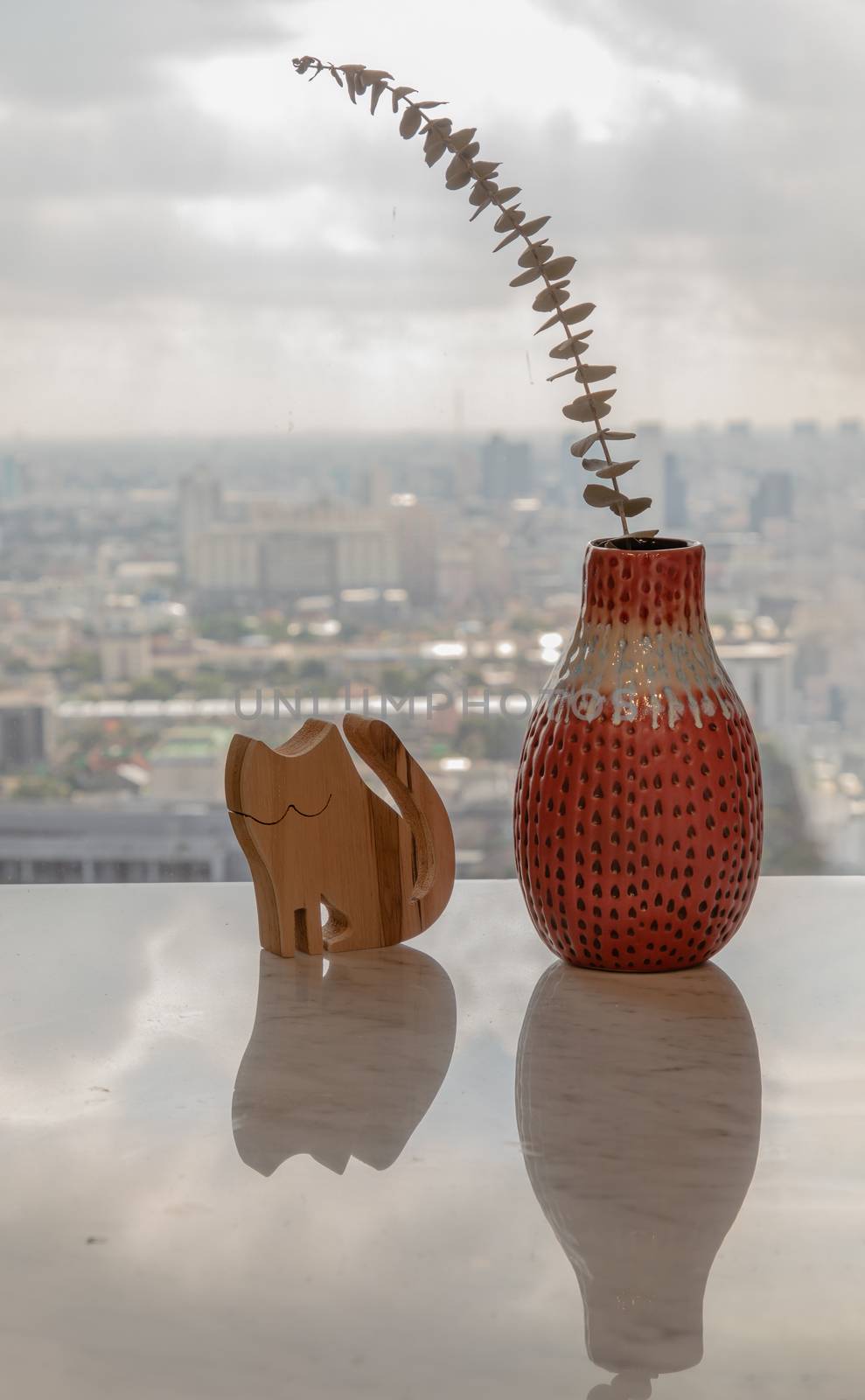 Dried flowers in beautiful ceramic red vase handmade with wooden doll in cat shape on marble table of living room and city view. Home decor. Space for text. Selective focus.