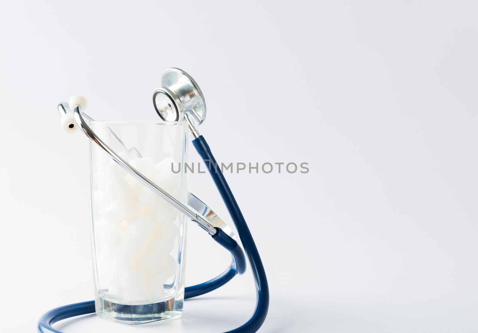 A glass full of white sugar cube sweet food ingredient and doctor stethoscope, studio shot isolated white background, health high blood risk of diabetes and calorie intake concept and unhealthy drink