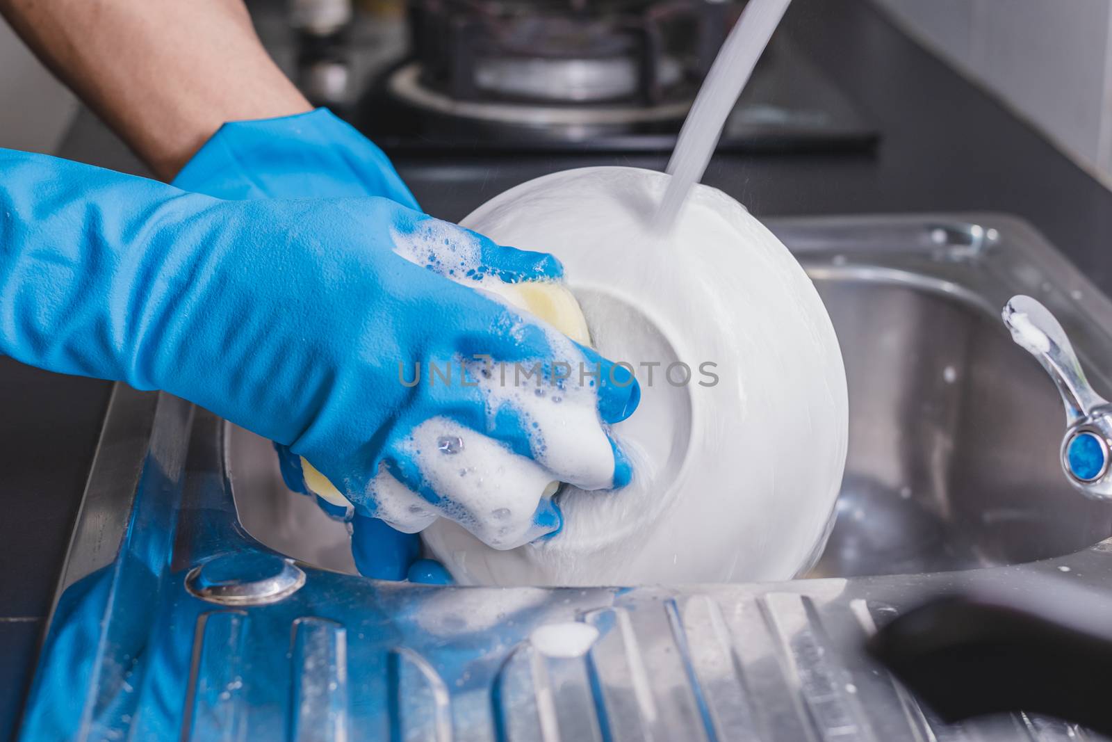 Close up of a man wearing a blue rubber glove was washing the cup with a dish washing liquid in the sink in the kitchen.