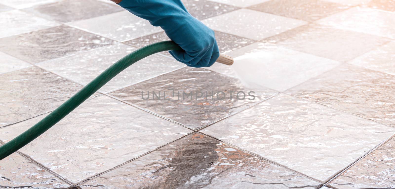 Hand of man wearing blue rubber gloves using a hose to cleaning the tile floor.