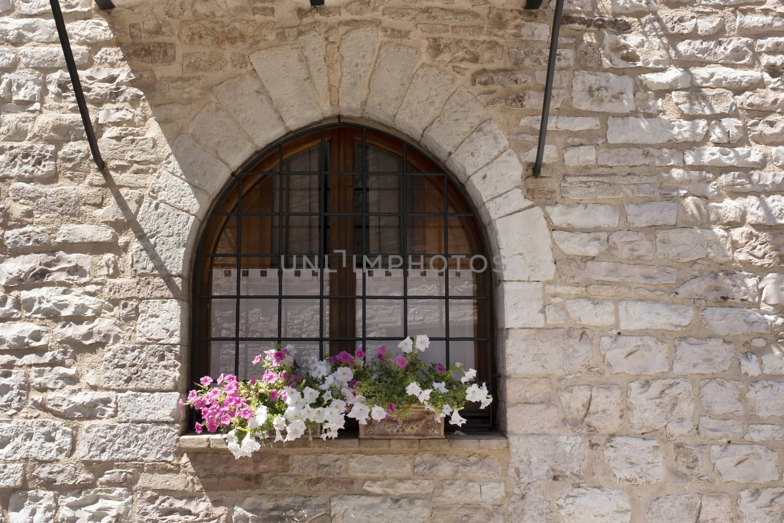A basket of bright pink and red flowers hangs on the window of a home in an ancient building in Italy by Tjeerdkruse