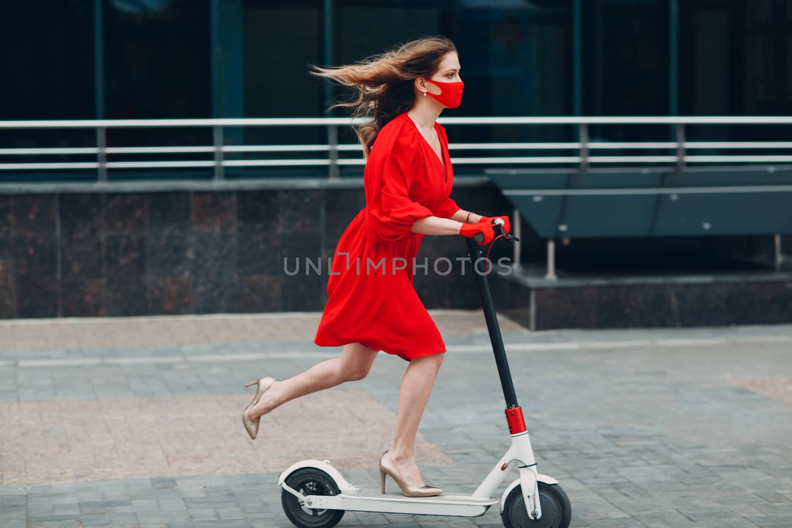 Young woman with electric scooter in red dress and gloves with face mask at the city. New normal fashion and coronavirus COVID protection concept