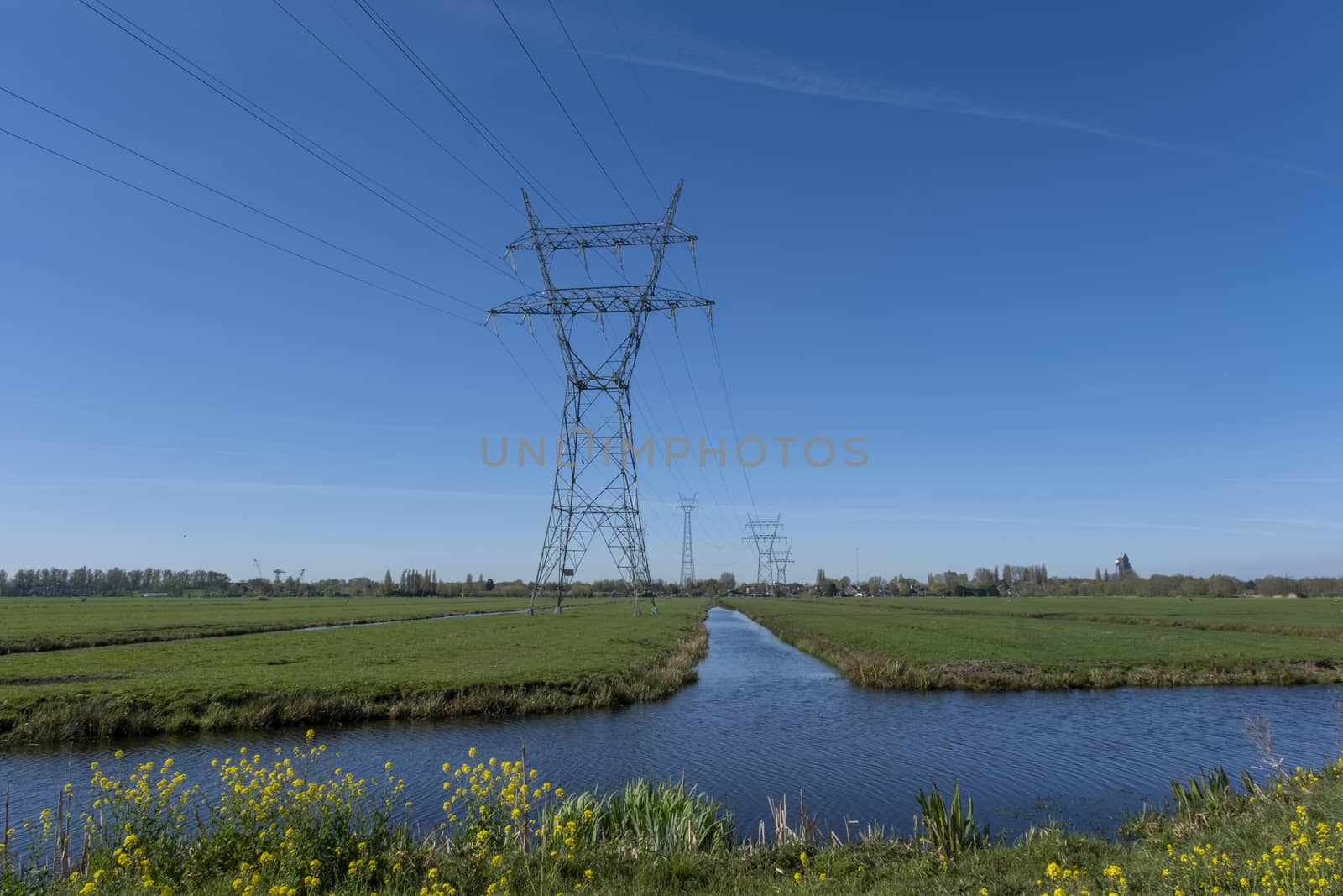 High voltage lines and power pylons in a flat and green agricultural landscape on a sunny day with cirrus clouds in the blue sky