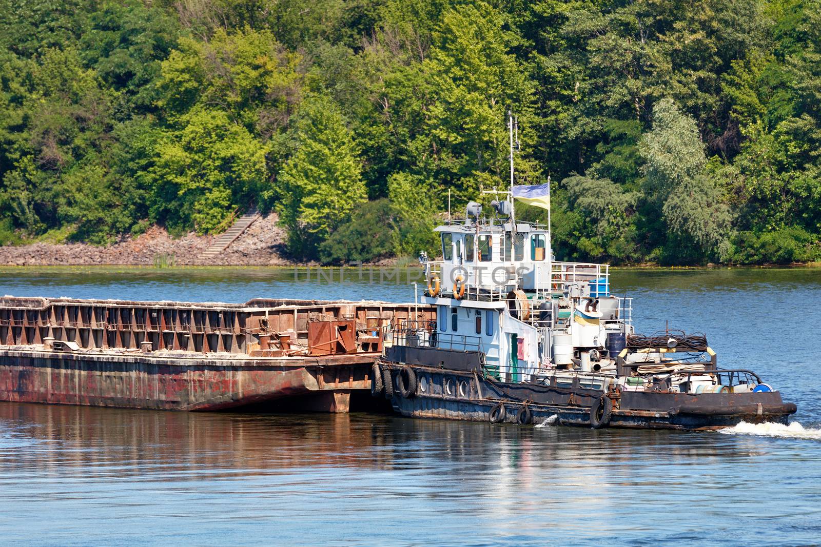 River tug slowly pushing an empty rusty barge down the river against coastal greenery, river freight transport concept, copy space.