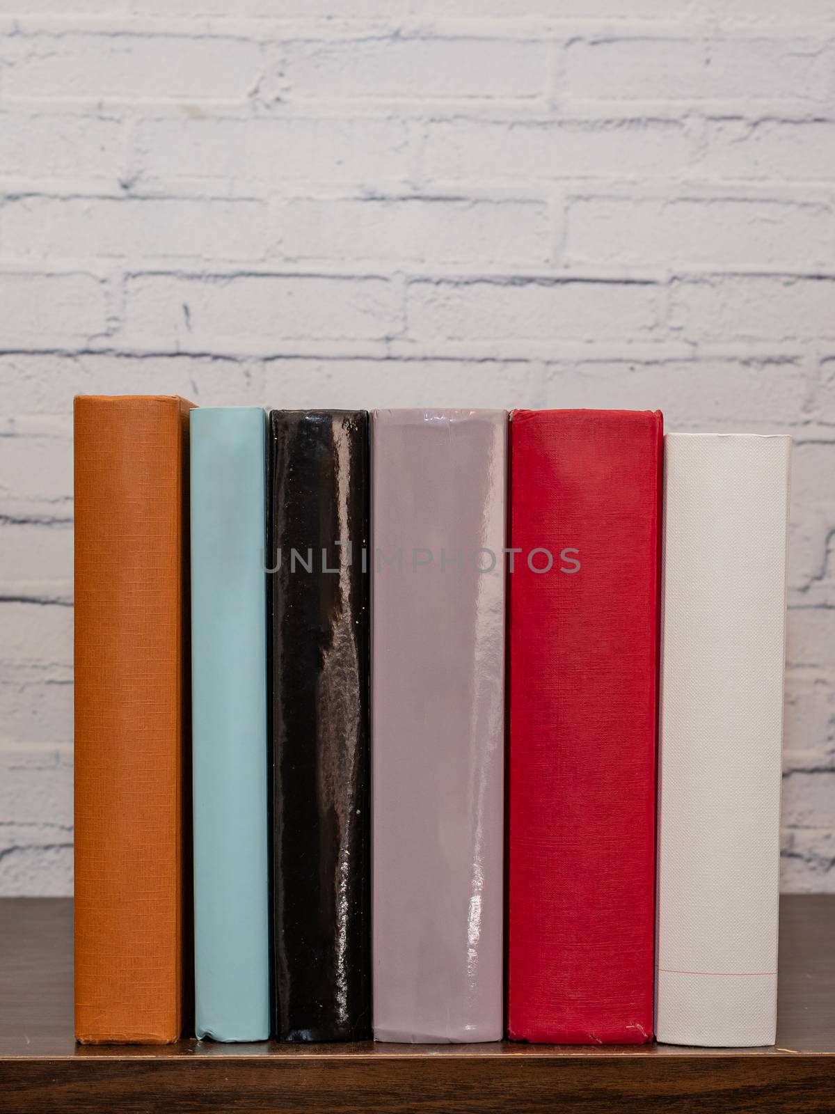 Six books of different colors resting on a dark wooden shelf, in the background a white painted brick wall