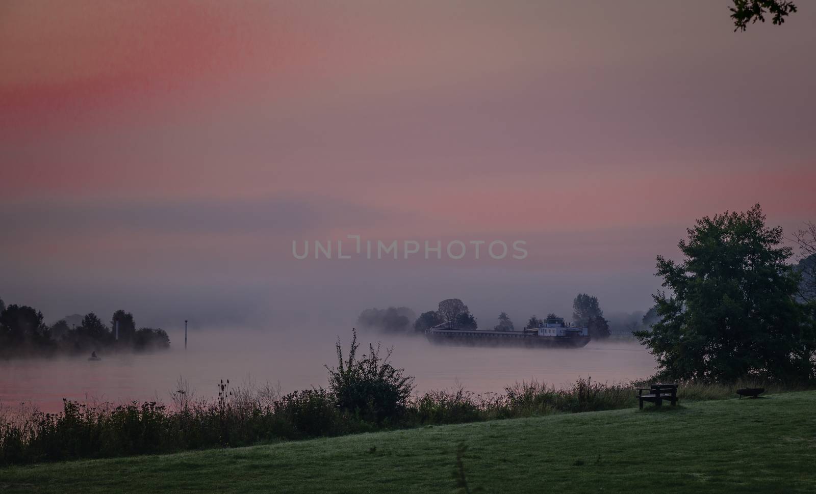 ship in the maas during sunrise in the early morning in limburg in holland with the trees mist and hazy fog