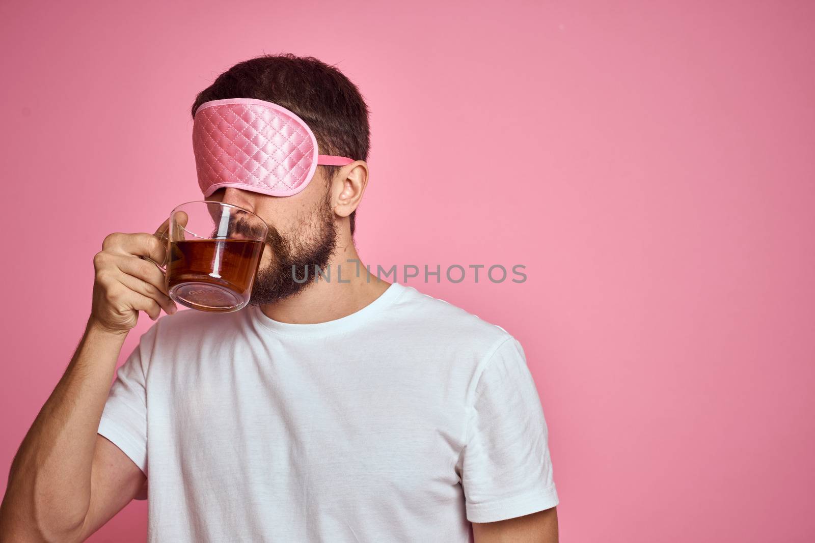 A man in a pink sleep mask with a cup of tea in his hands on an isolated background cropped view by SHOTPRIME