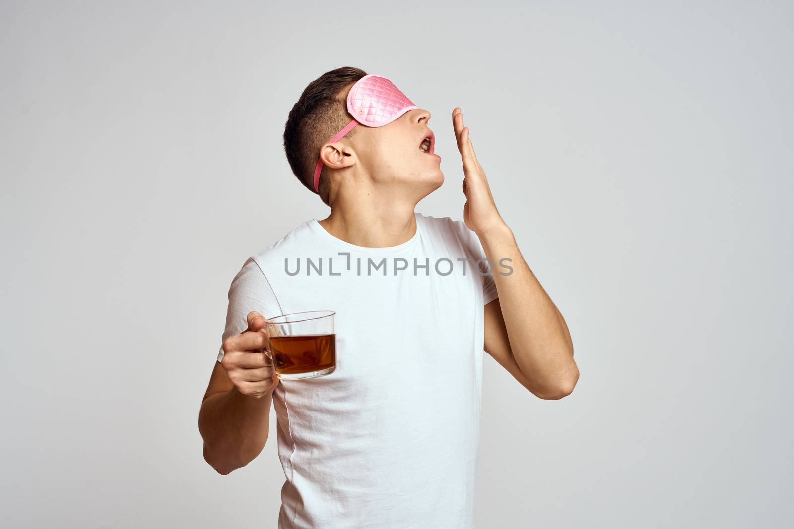 handsome man with pink sleep mask and with a cup of tea in hand pulls himself up on a light background by SHOTPRIME