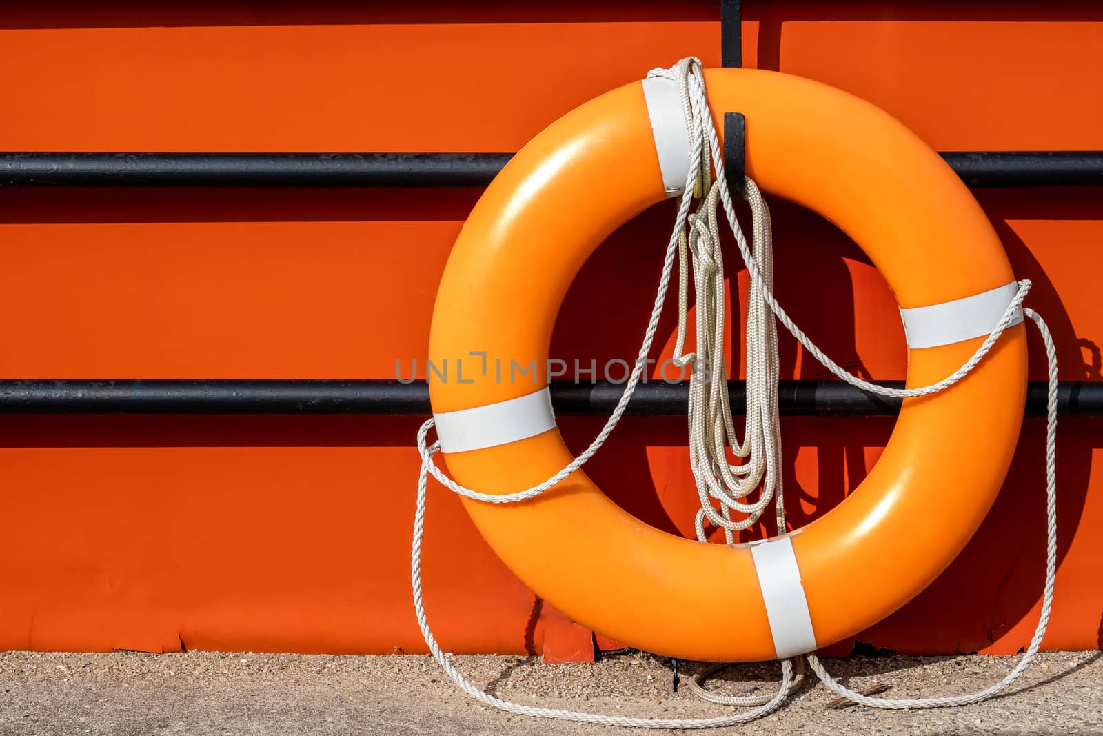 An orange life ring hangs on a hook on a black fence in front of an orange background surface. The life preserver has white ropes attached.