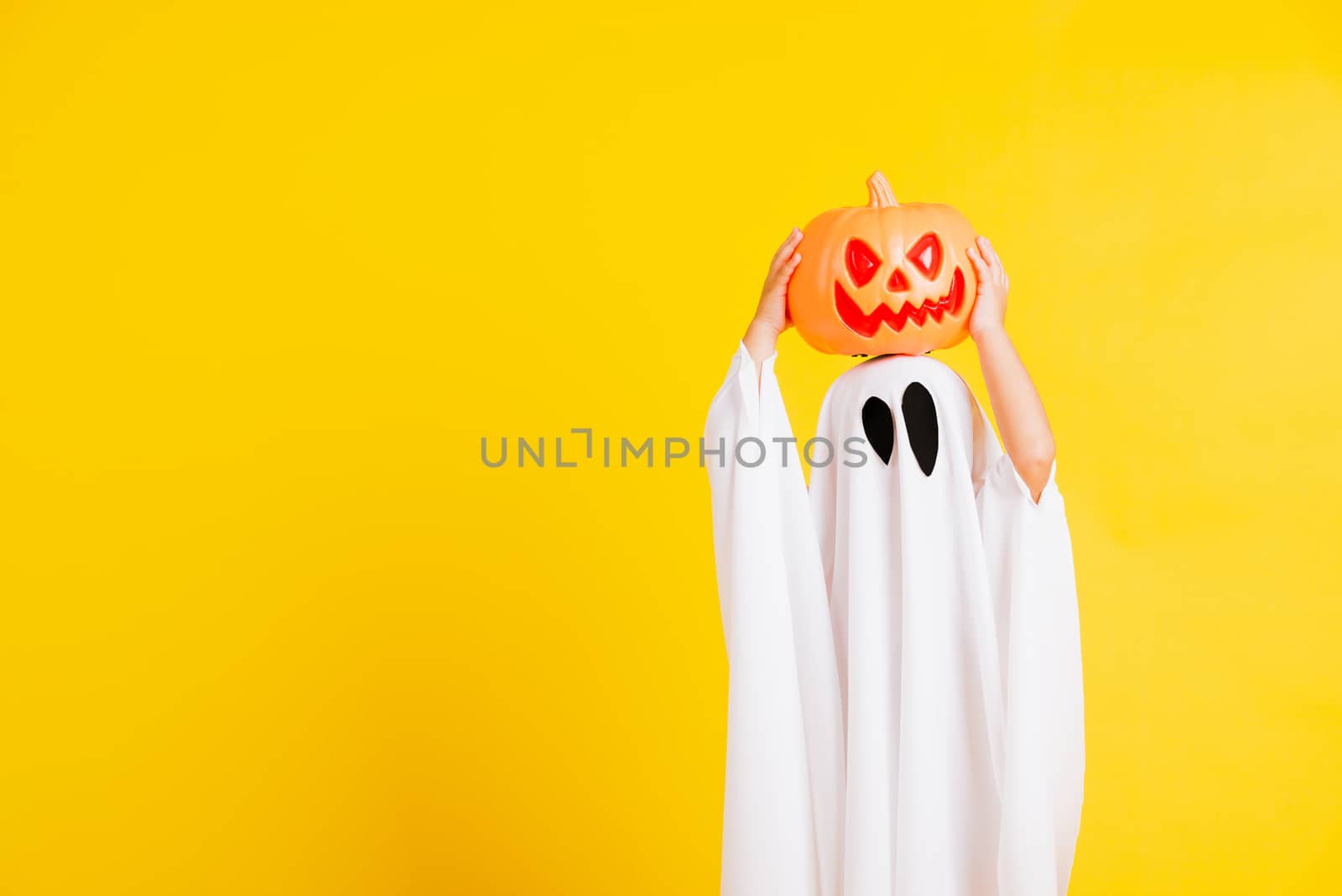 Funny Halloween Kid Concept, little cute child with white dressed costume halloween ghost scary he holding orange pumpkin ghost on hand, studio shot yellow on white background