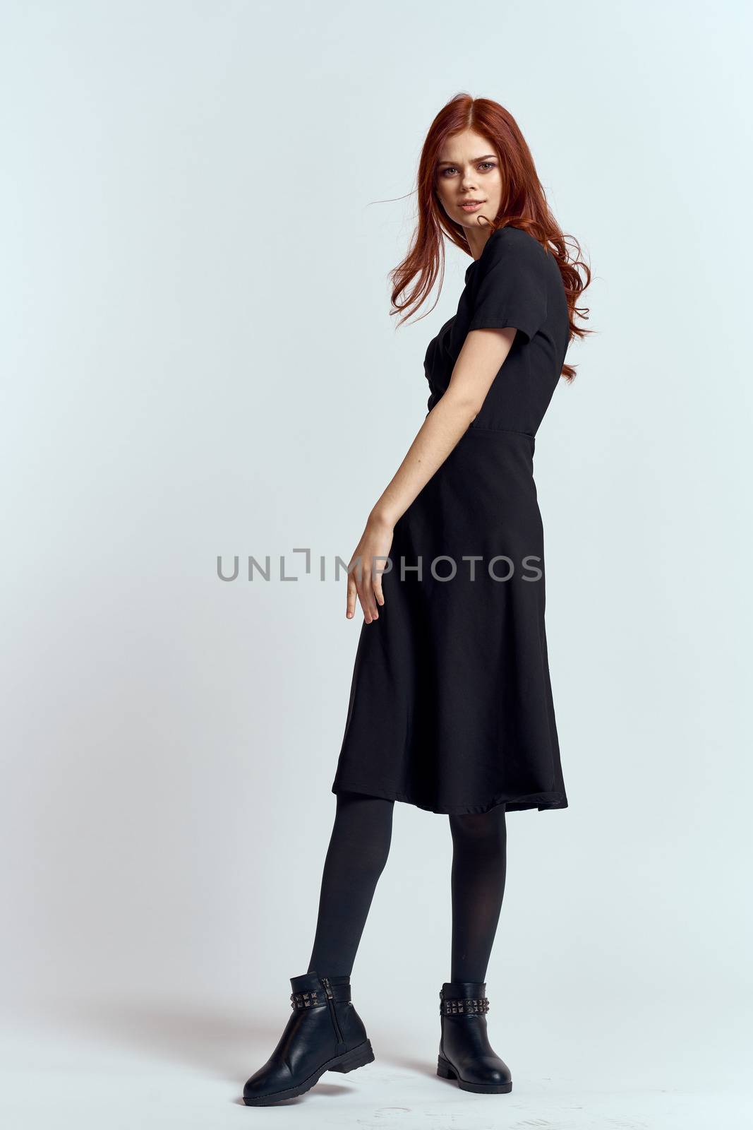 A woman in a black dress on a light background and pantyhose shoes red hair and pose in full growth. High quality photo