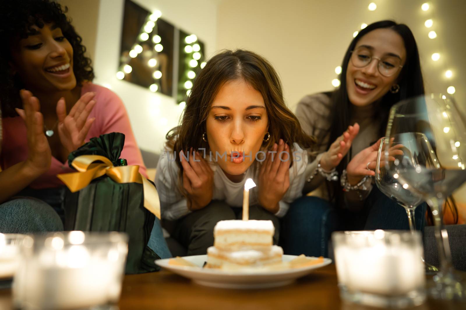 Close up of cute young woman blows on the cake candle next to her present celebrating her birthday at home with her two mixed race hispanic and caucasian female best friends by robbyfontanesi