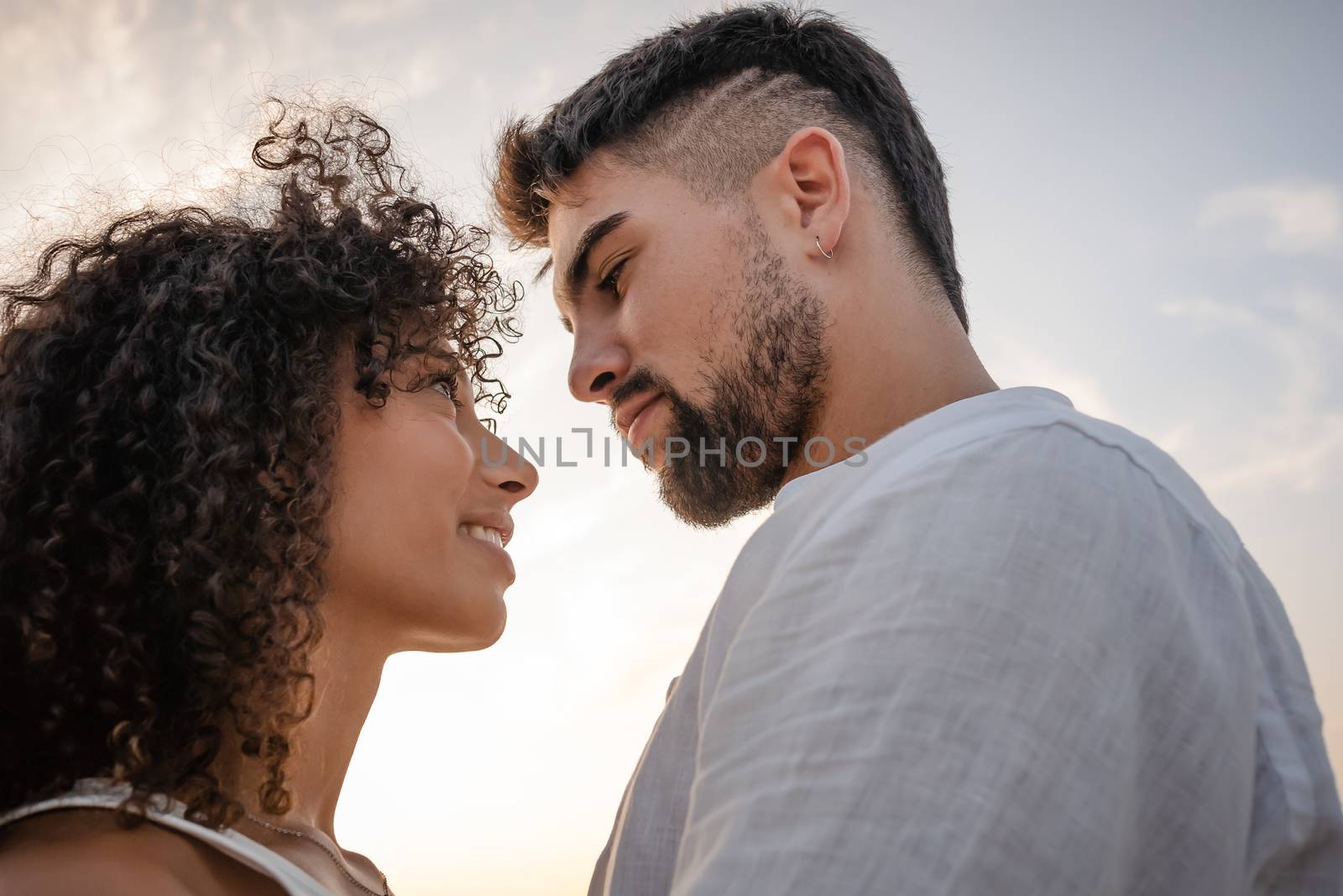 Side view of two young multiethnic passionate lovers looking in the eyes each other - Macho bearded sexy man looks at his Latin woman with ardor in a romance sunset scene ideal for a book cover by robbyfontanesi