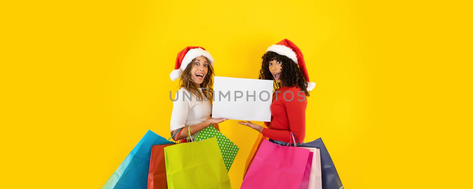 Two young cute women wearing Santa Claus hat looking to the camera with astonished face, open mouth, wide eyes, holding colored shopping bags - Christmas banner with yellow background for copy space by robbyfontanesi