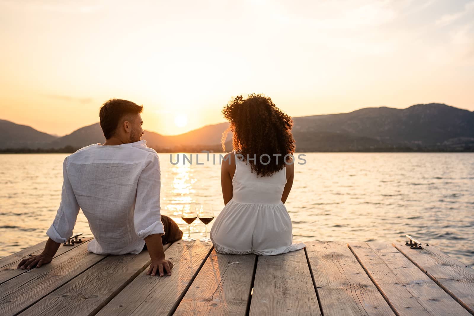 Multiracial young couple caucasian and hispanic sitting on a jetty with two red wine glasses and setting sunlight between them - Romantic sunset scene with light reflection on the water and lovers