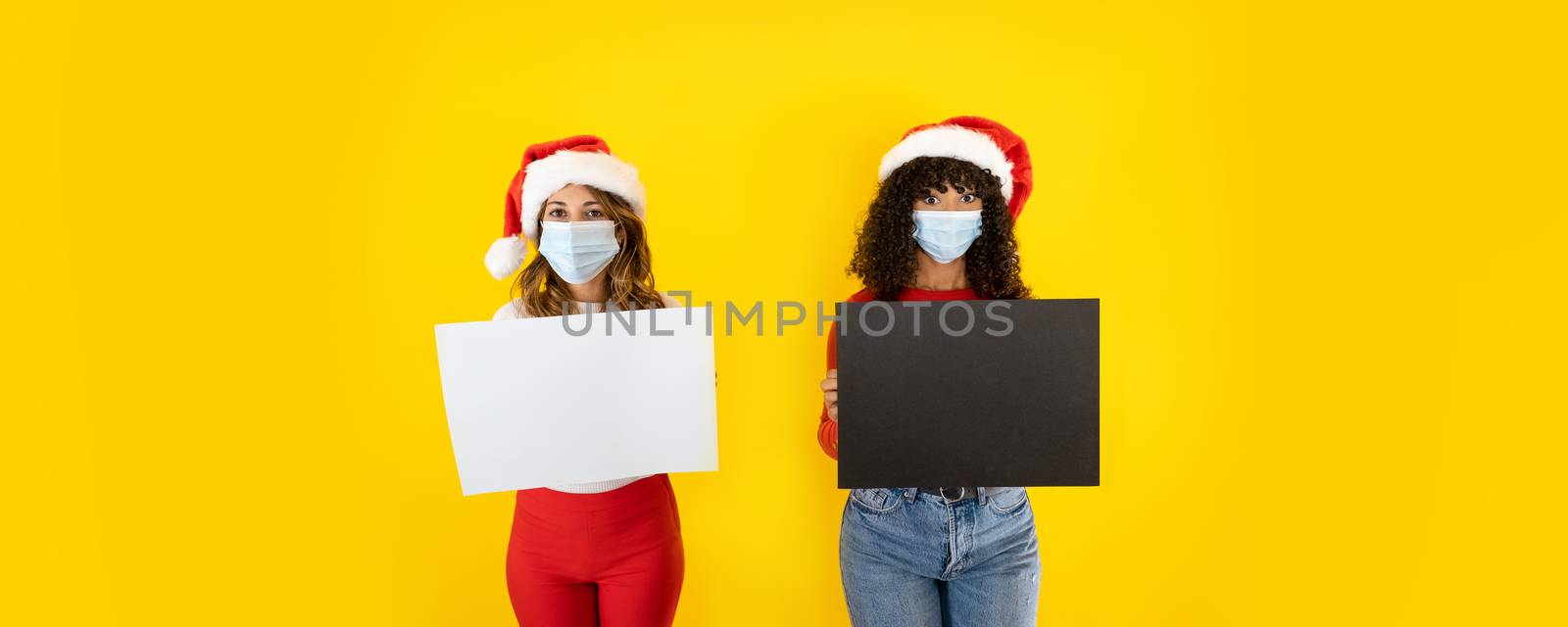 Two young cute women wearing Santa Claus hat looking to the camera with wide eyes and medical mask, holding sign for copy space - Christmas banner for Coronavirus by robbyfontanesi