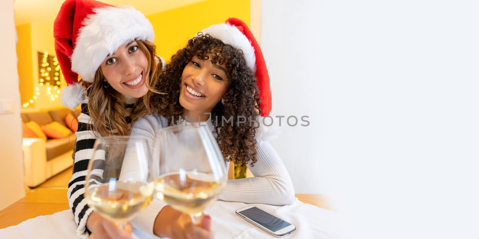 Two mixed race Caucasian and Hispanic fine young women at home looking at the camera for toasting with a glass of wine in video call - New normal holidays celebration for Coronavirus outbreak by robbyfontanesi
