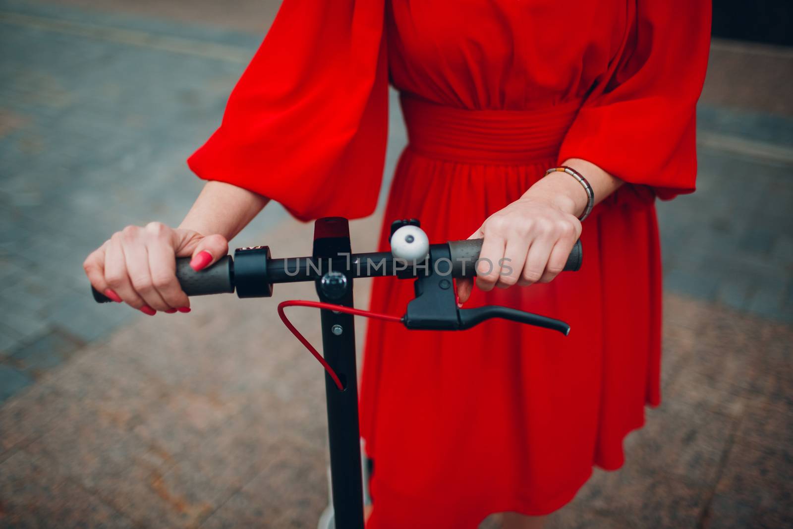 Young woman holds steering wheel of electric scooter in red dress at the city.