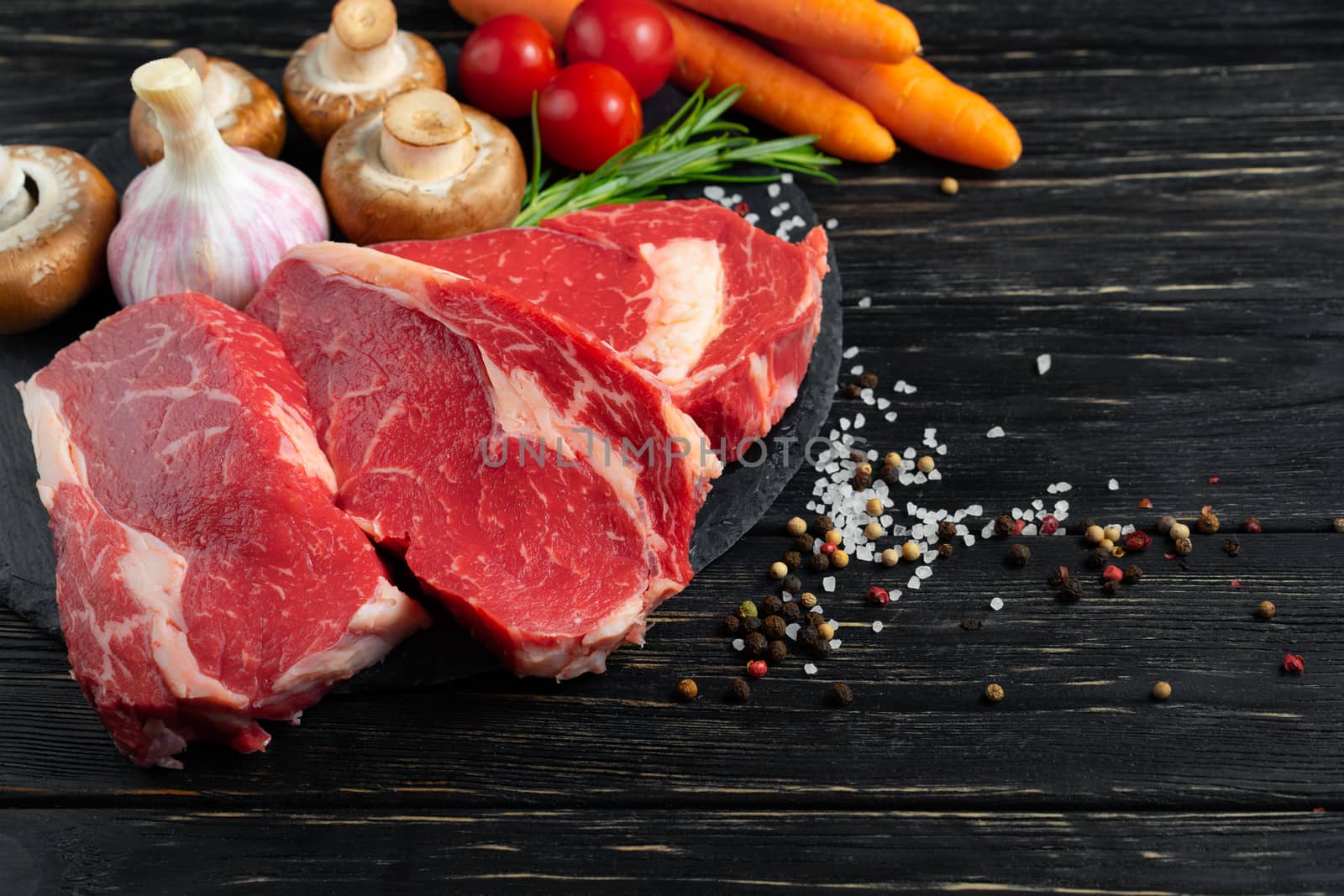 Three pieces of juicy raw beef with rosemary on a stone cutting board on a black wooden table background. Meat for barbecue or grill with vegetables pepper and salt seasoning