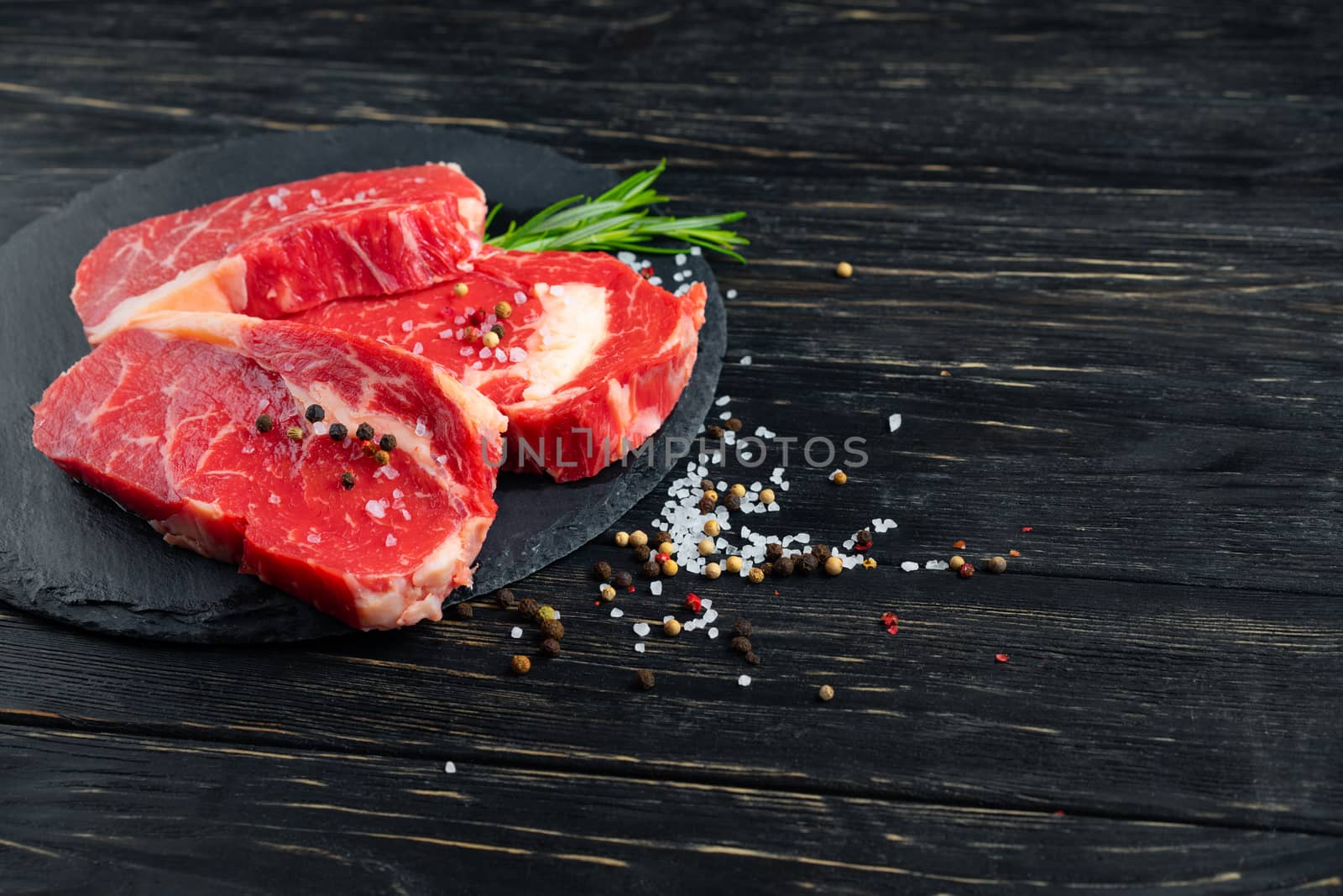 Three pieces of juicy raw beef with rosemary on a stone cutting board on a black wooden table background. Meat for barbecue or grill sprinkled with pepper and salt seasoning