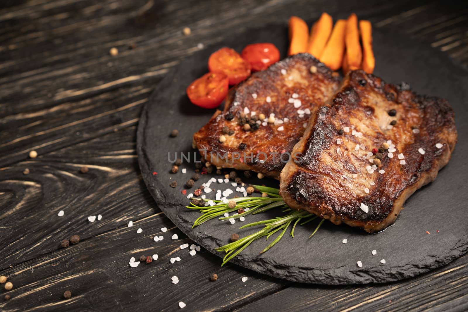 A juicy piece of fried meat with grilled cherry tomatoes and carrot lies on a stone plate against a black wooden table. Degree of roasting well done. Food concept with copy space