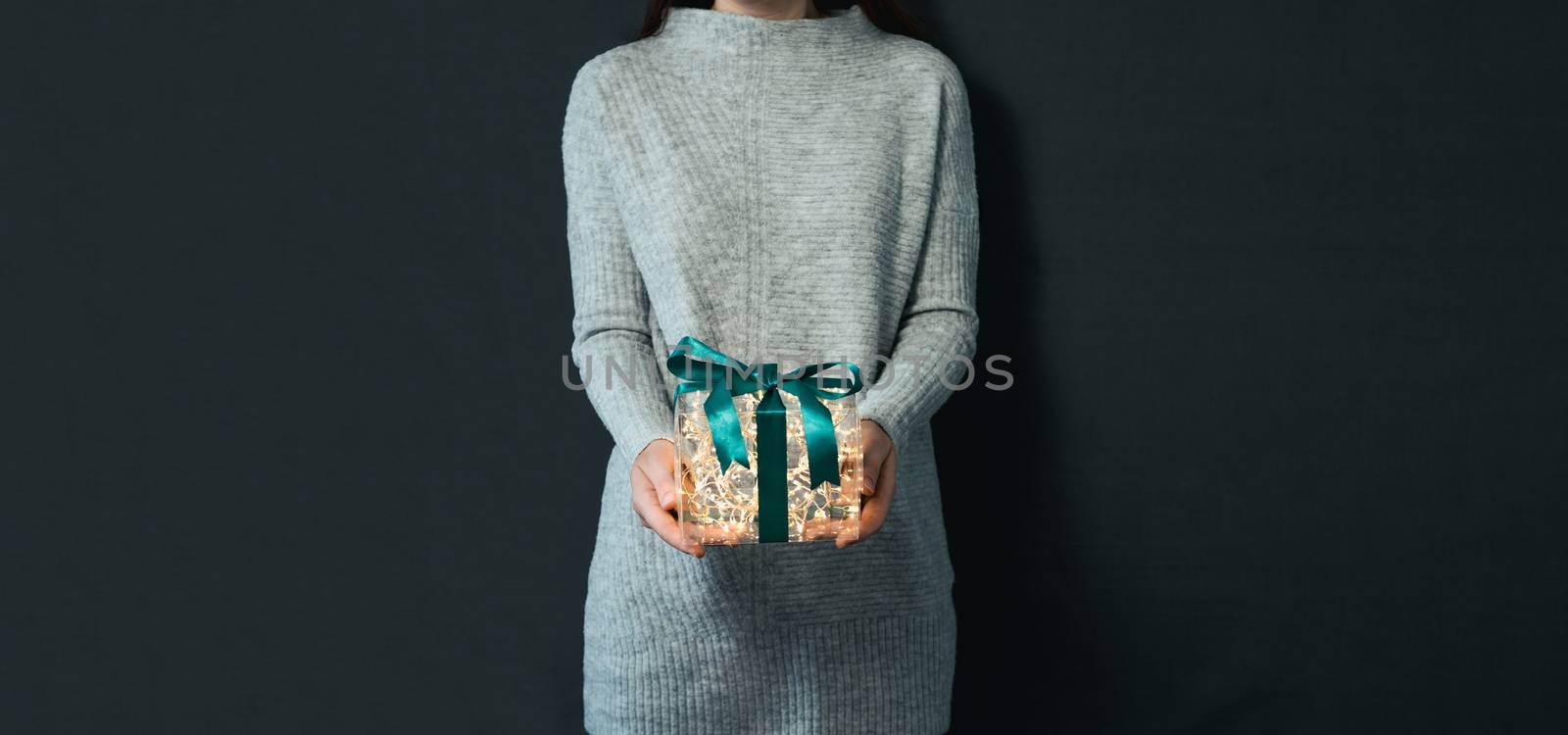 Woman in gray pullover holding luminous gift box in hands on dark background. Glowing yellow light bulbs into transparent gift box with dark green ribbon. Christmas and New Year concept