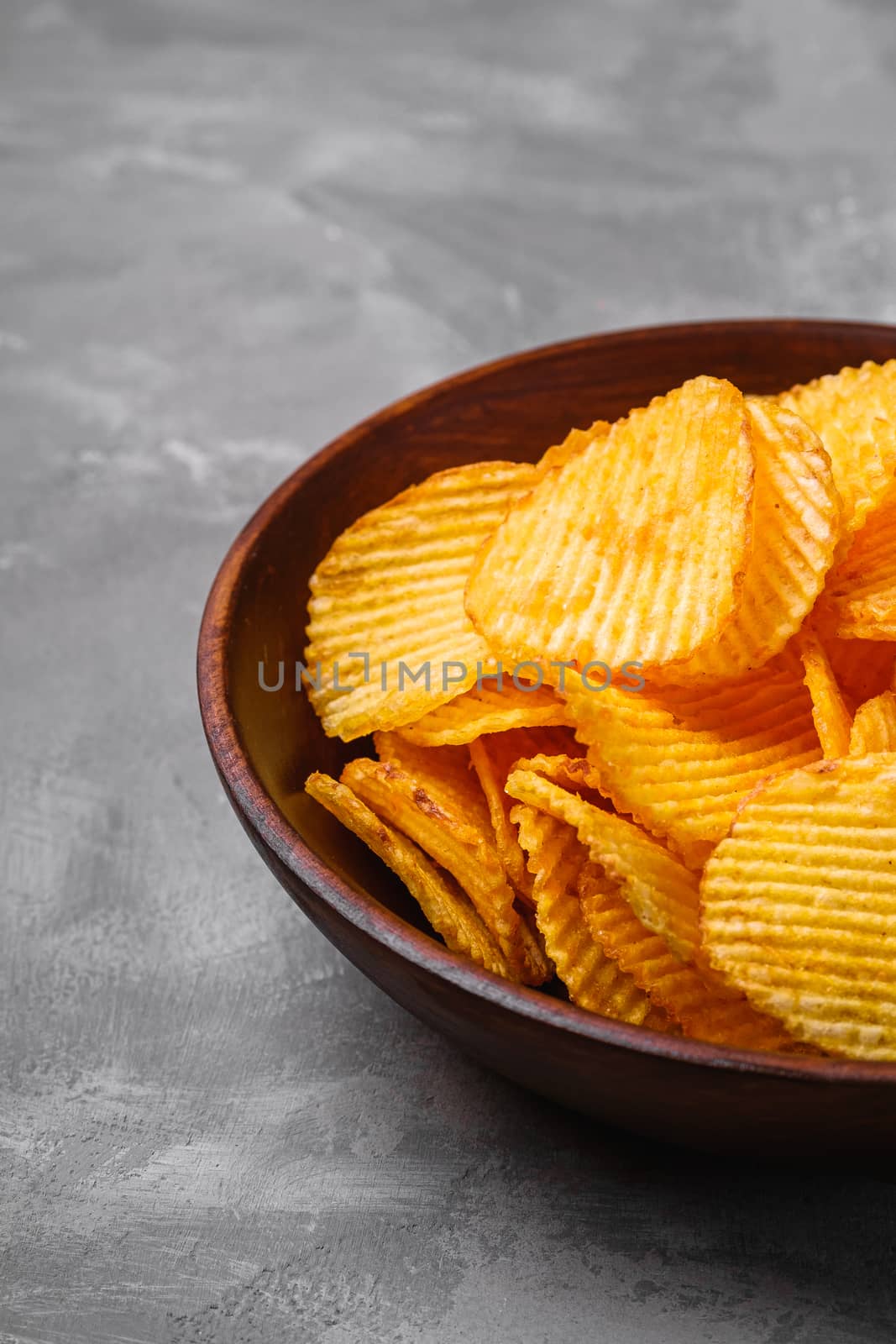 Fried corrugated golden potato chips in brown wooden bowl on concrete background, angle view