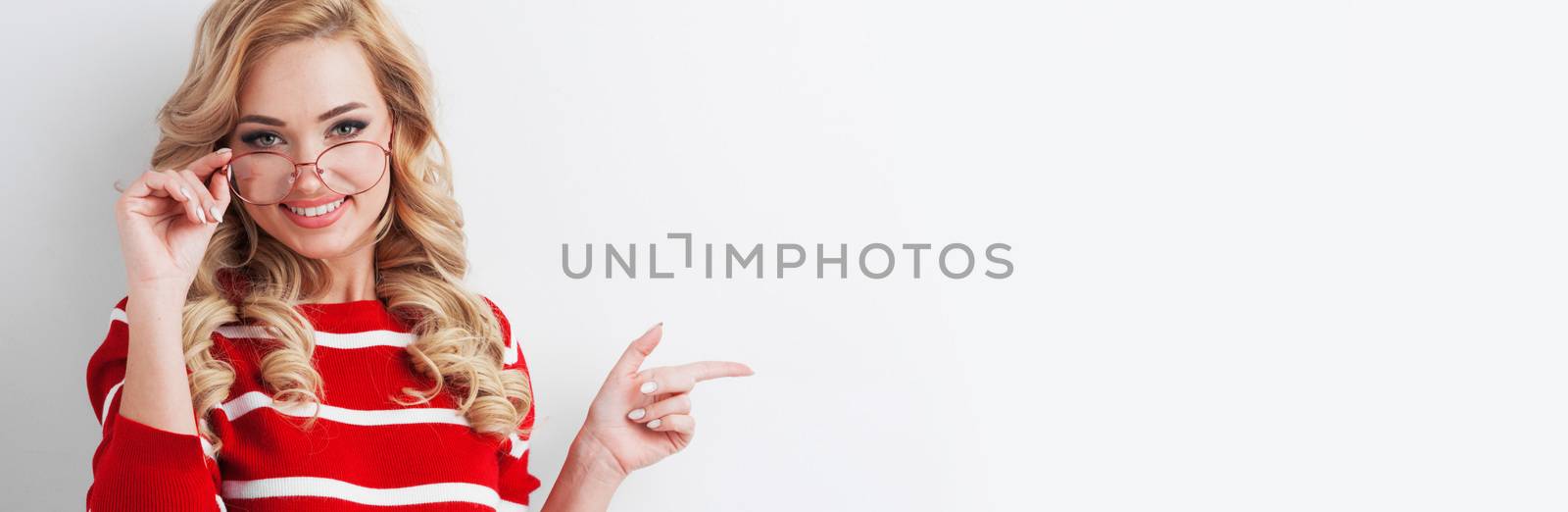 Cheerful Caucasian blonde female with glad expression, indicates left with positive expression, pointing aside, happy to advertise something on blank white background