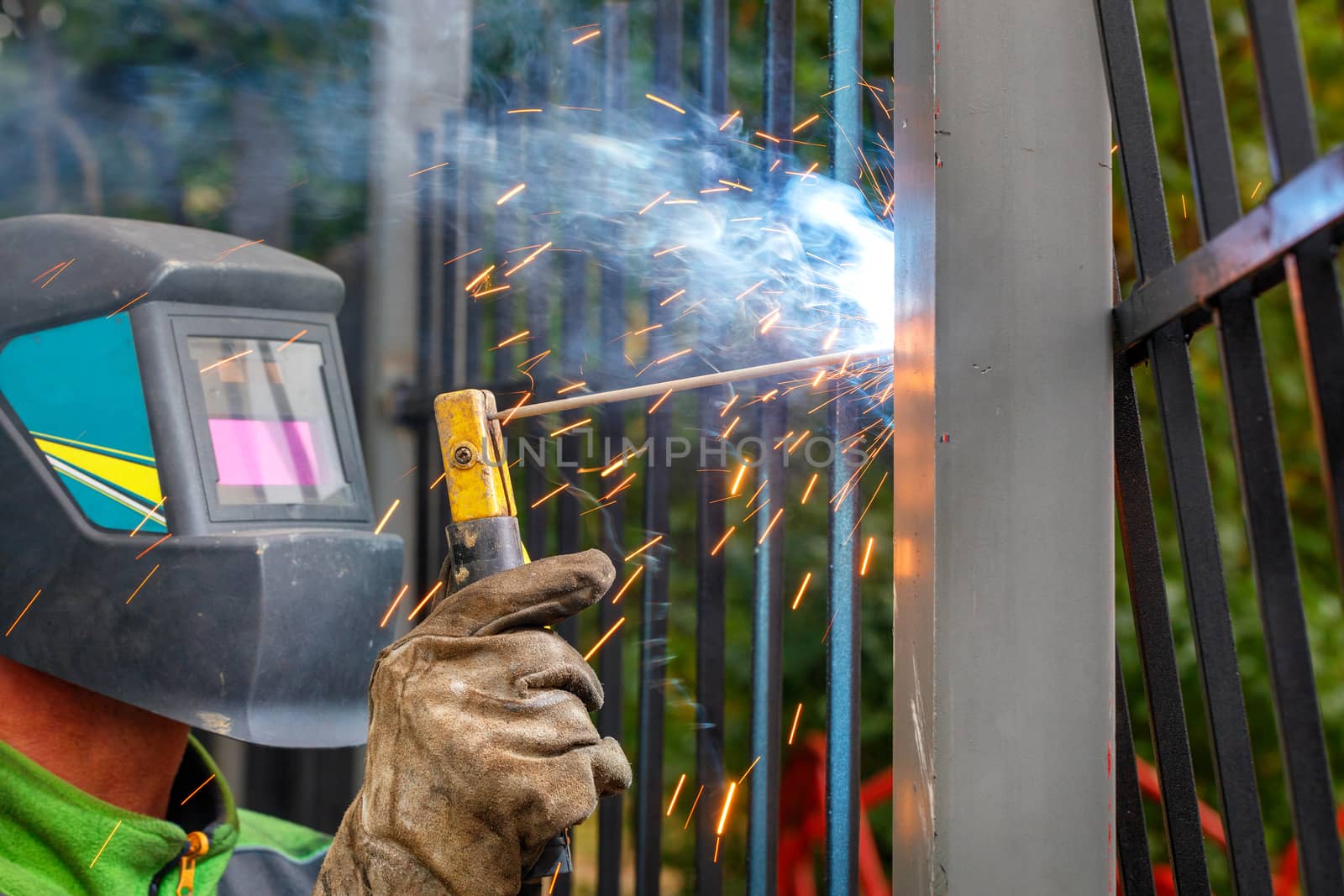A welder in a protective helmet and gloves using an electrode welds a metal fence in a park area, bright sparks, blue smoke fly, selective focus, copy space.