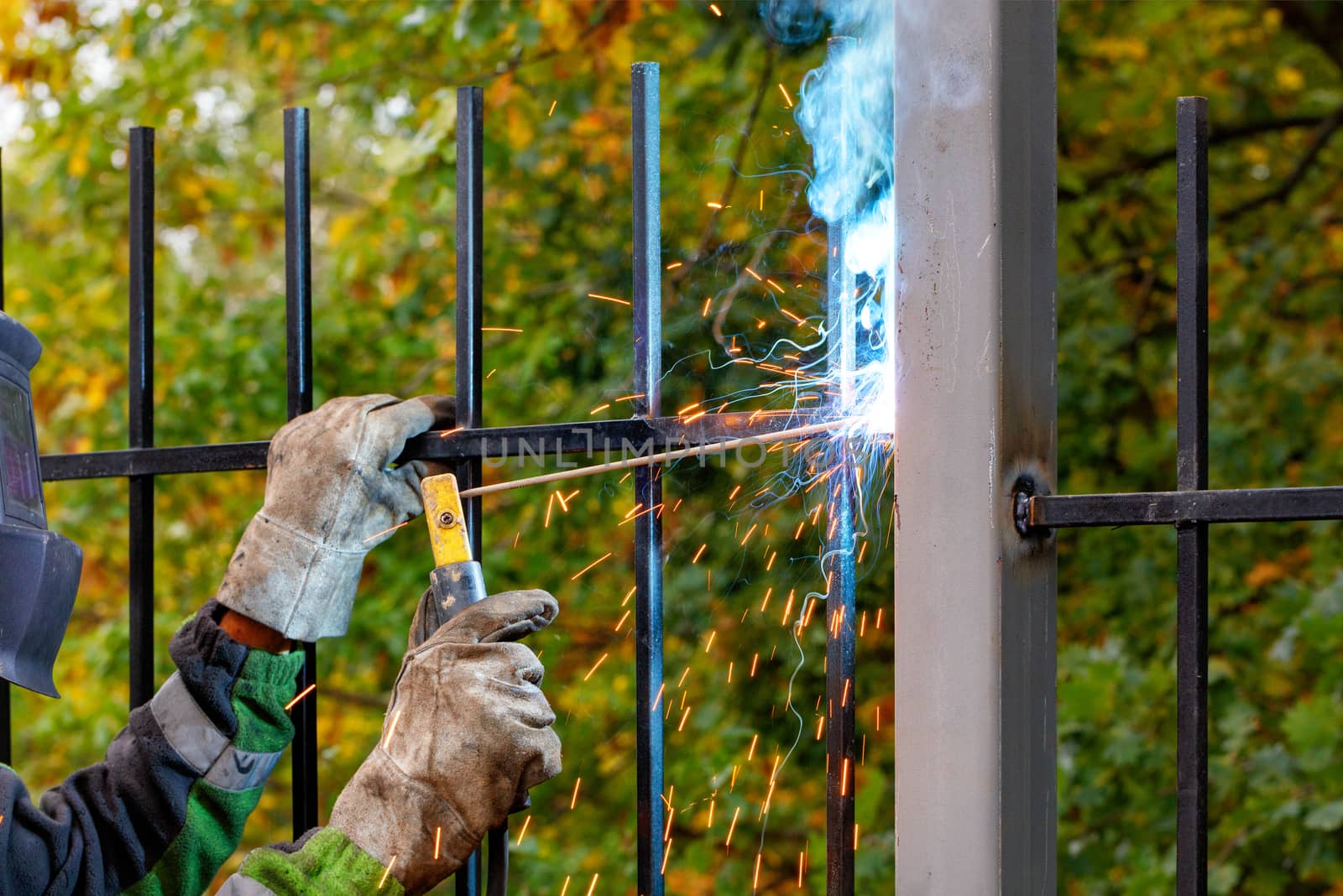 A welder in a protective helmet and gloves uses an electrode to weld a metal fence in a park area, against a blurred background of autumn greenery. Selective focus, copy space.