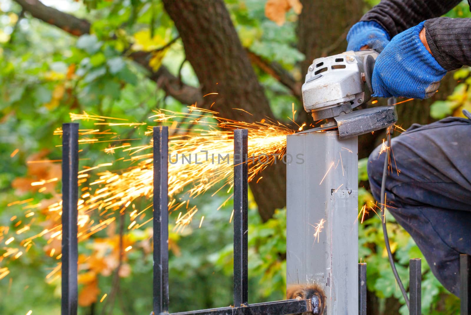An industrial worker cuts a metal post with a disc angle grinder, creating a bright plume of many hot sparks.