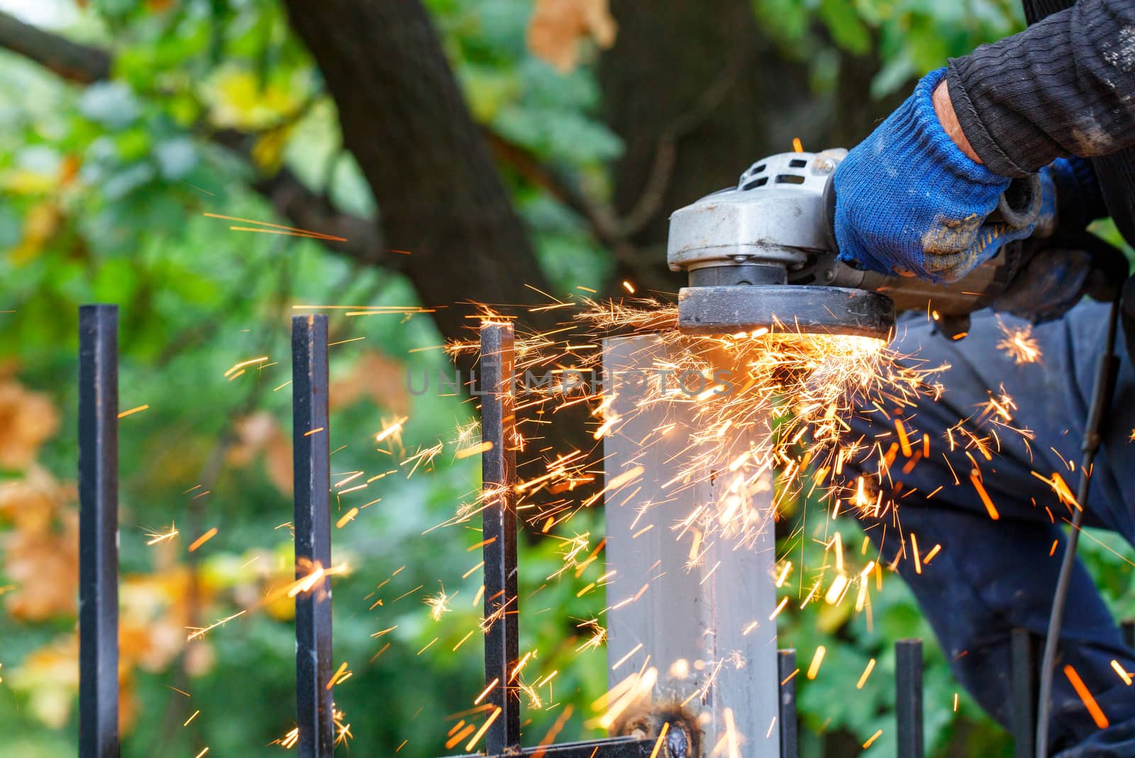 A bright plume of many hot sparks scatters as an artisan works, cutting a metal post with a disc angle grinder against a blurred background of green park.