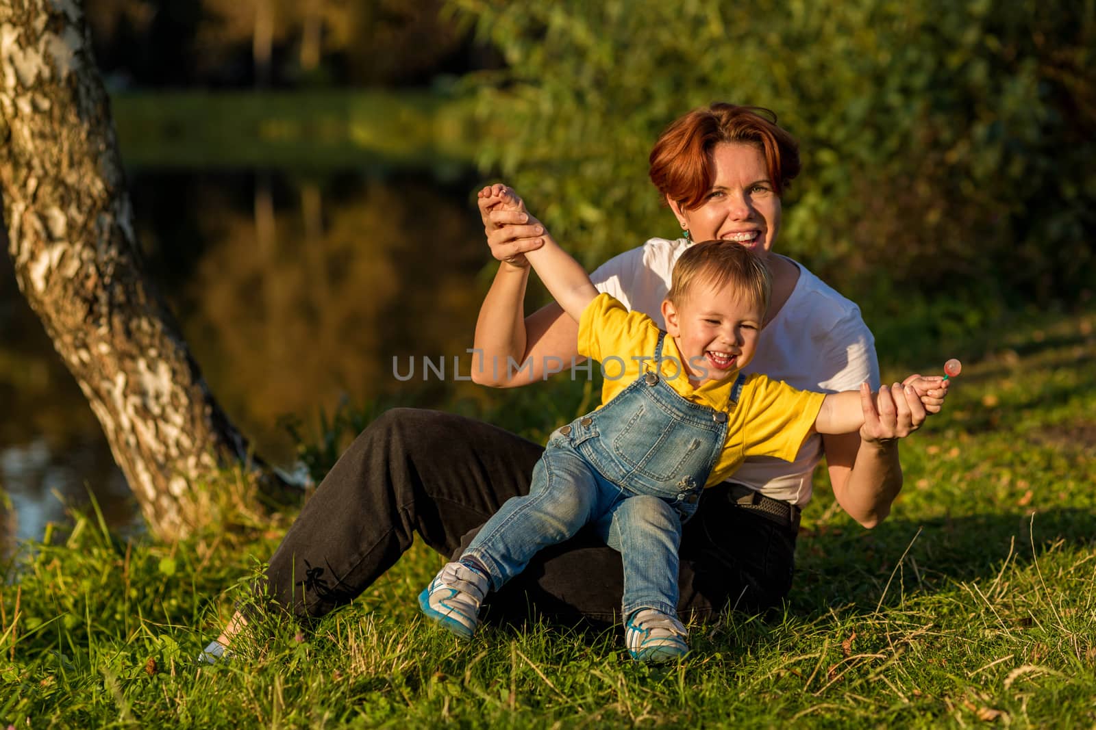 Mom and little boy sit on the river bank in the rays of the setting sun by galinasharapova