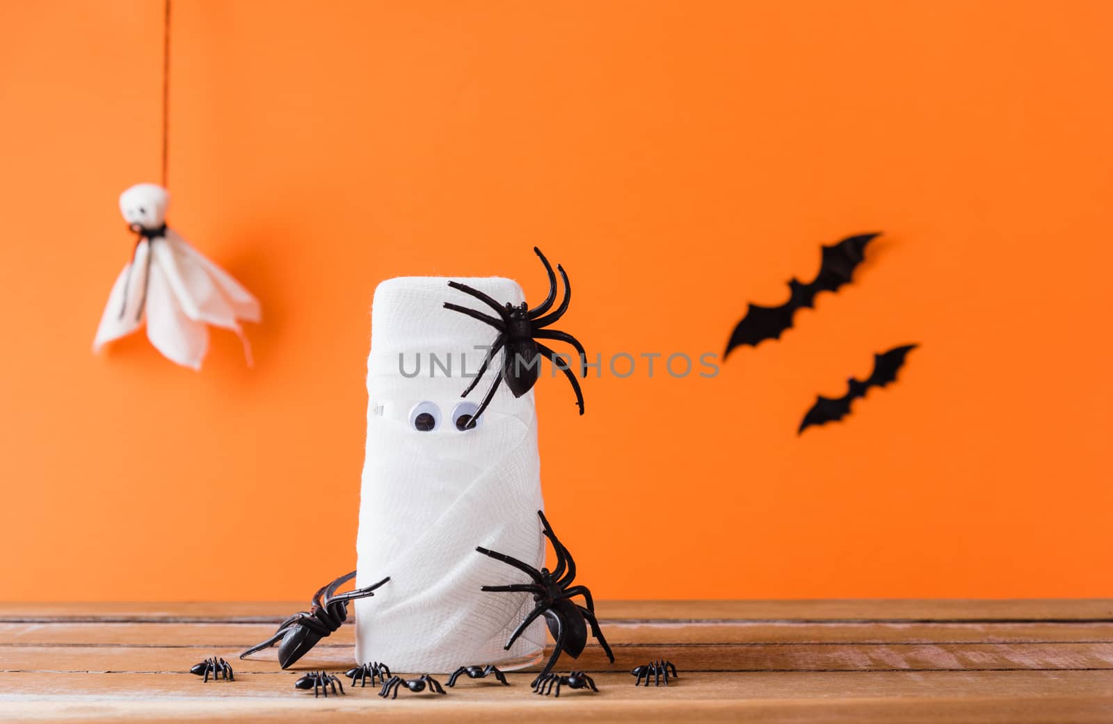 Funny Halloween day decor party concept, The mummy ghost on water glass wrapped around with bandage and have bats and spider stick it found only eyes, studio shot isolated on orange background
